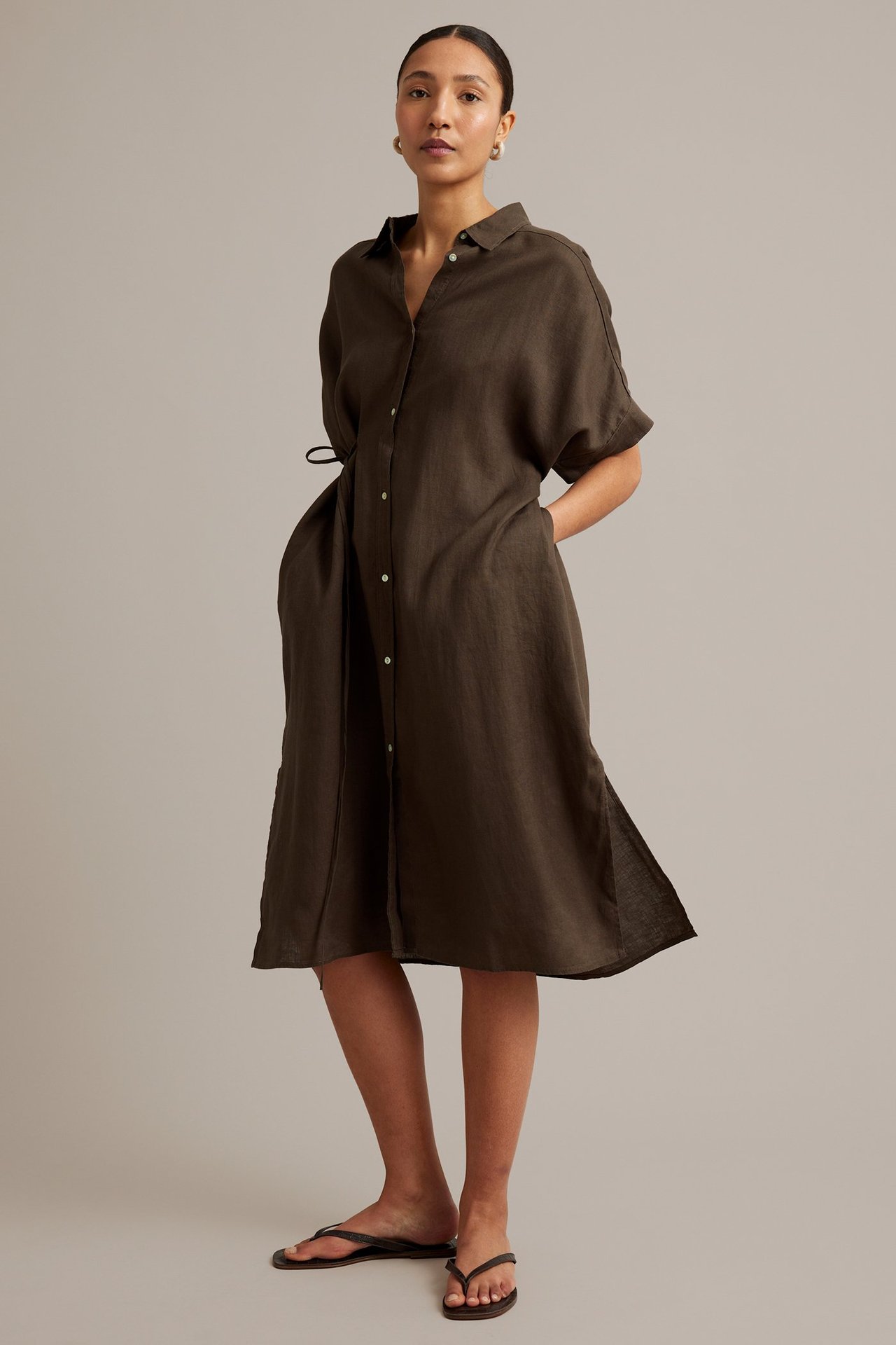 Kvinne i mørkbrun linskjortekjole med knapper, snøring i midjen, lommer, brune flip-flops og gulløredobber.