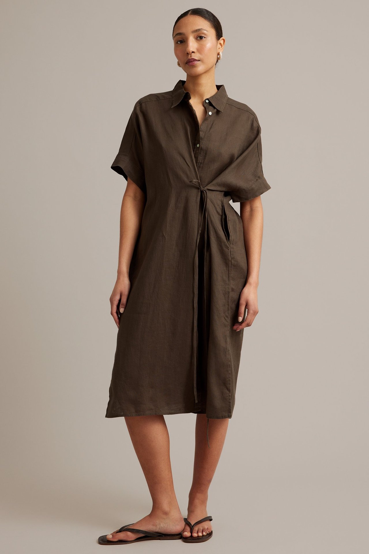 Frontbilde av en kvinne i brun skjortekjole av lin med knytmidje og mørke flip-flops.