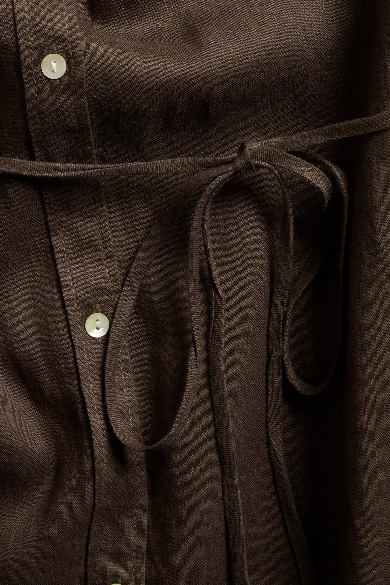Närbild av en brun linneskjortklänning med ljusa knappar och knuten midjesnodd.
