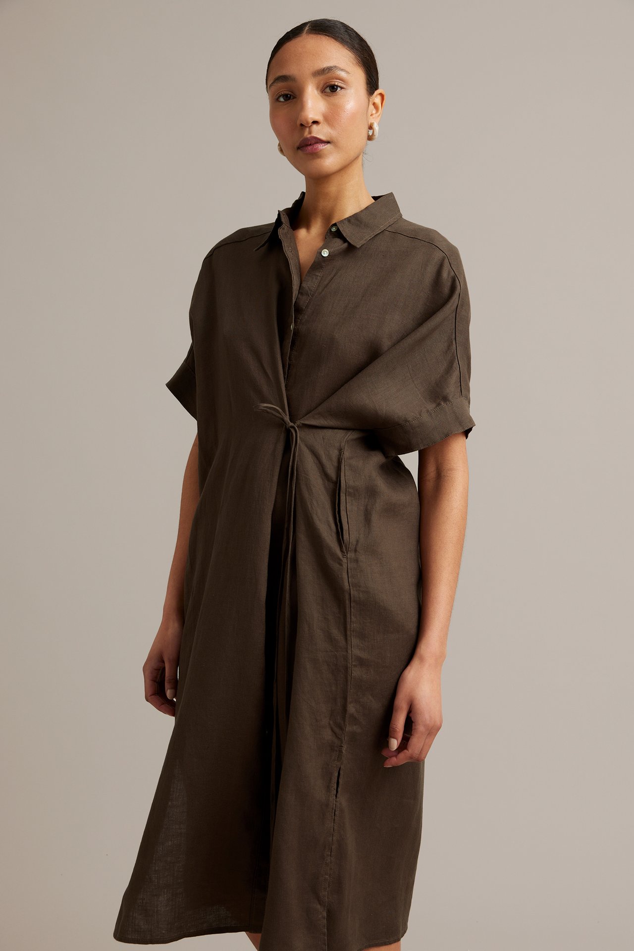 Framifrån: Kvinna i brun linneskjortklänning med knyt i midjan och korta ärmar, bär örhängen.