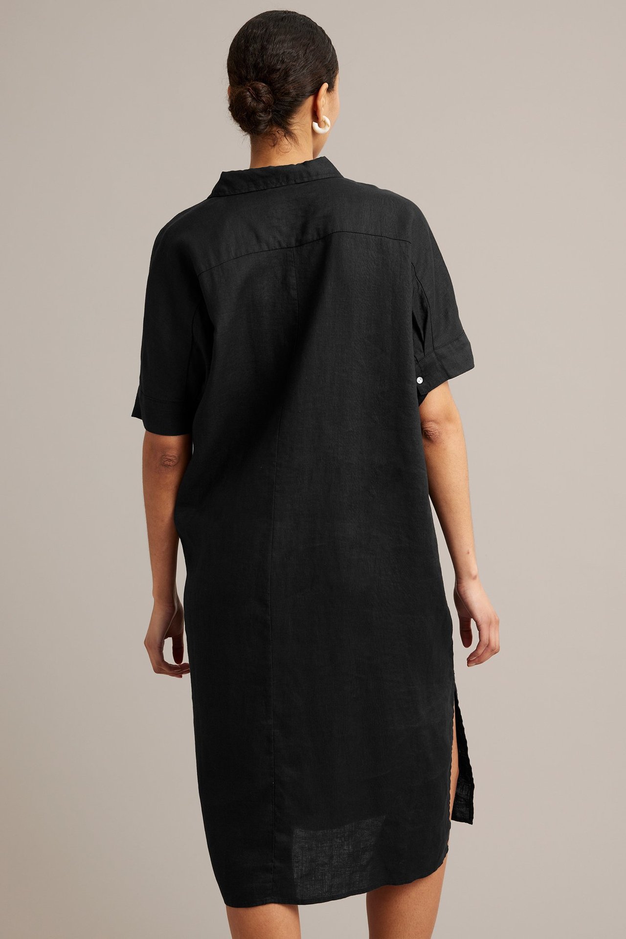 Ryggvisning av en kvinne i en svart skjortekjole i lin med korte ermer og hvite creoler.