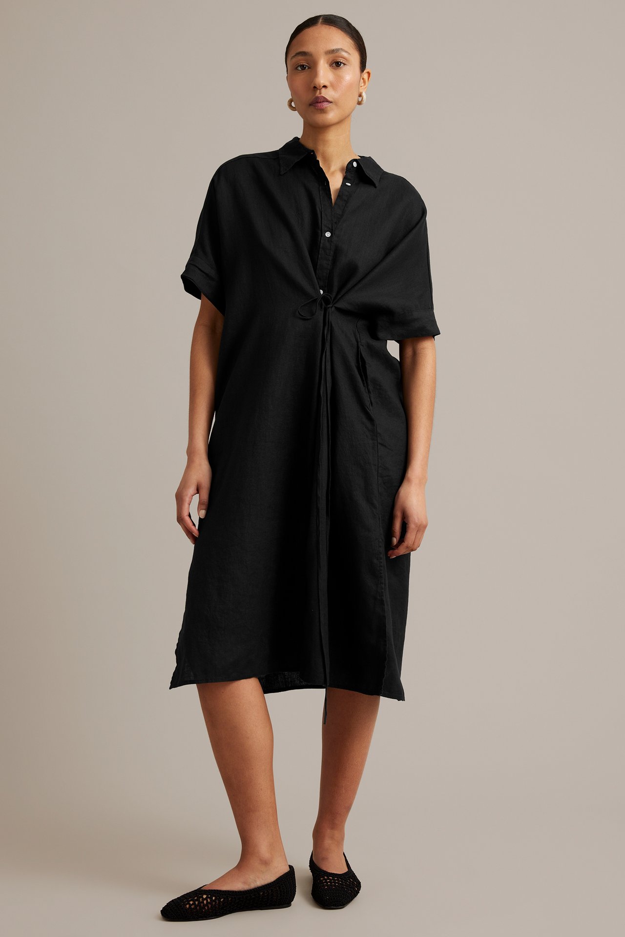 Kvinna, framifrån, i svart linneskjortklänning med dragsko i midjan och svarta vävda platta skor.