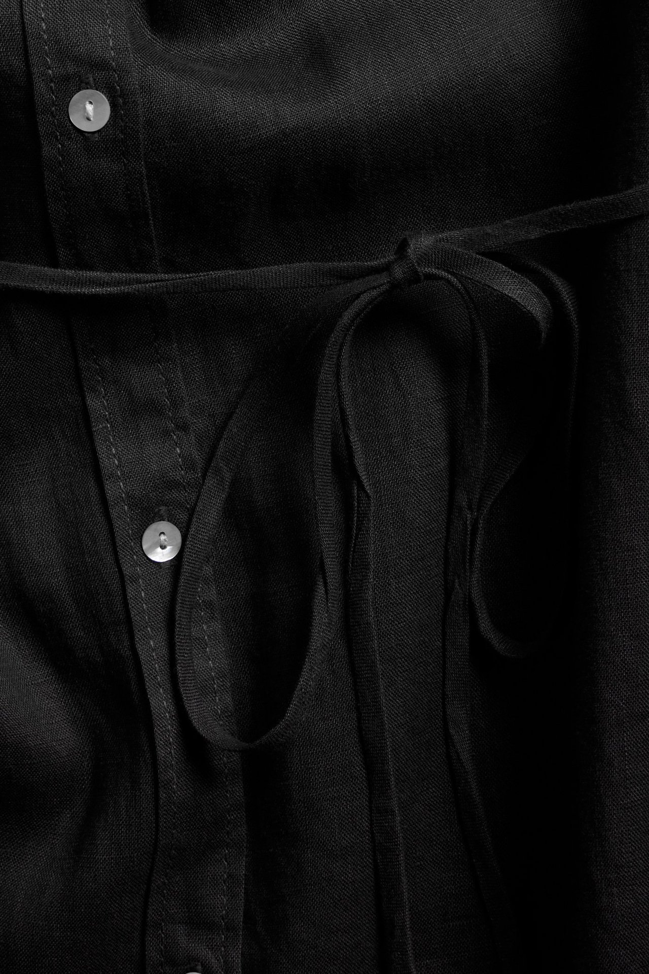 Närbild av en svart linneskjortklänning med pärlemorknappar och knutet midjeband.