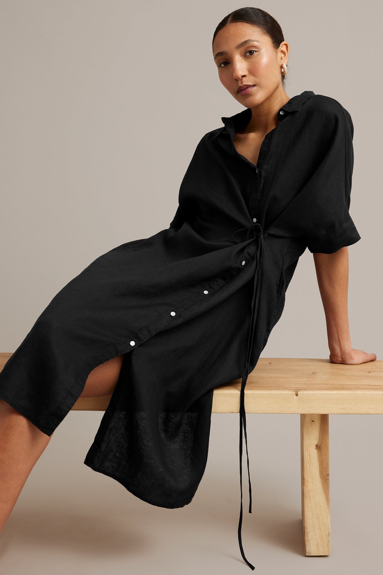 Sned frontvy av kvinna i svart linneskjortklänning med knytmidja, vita knappar, guldörhängen.