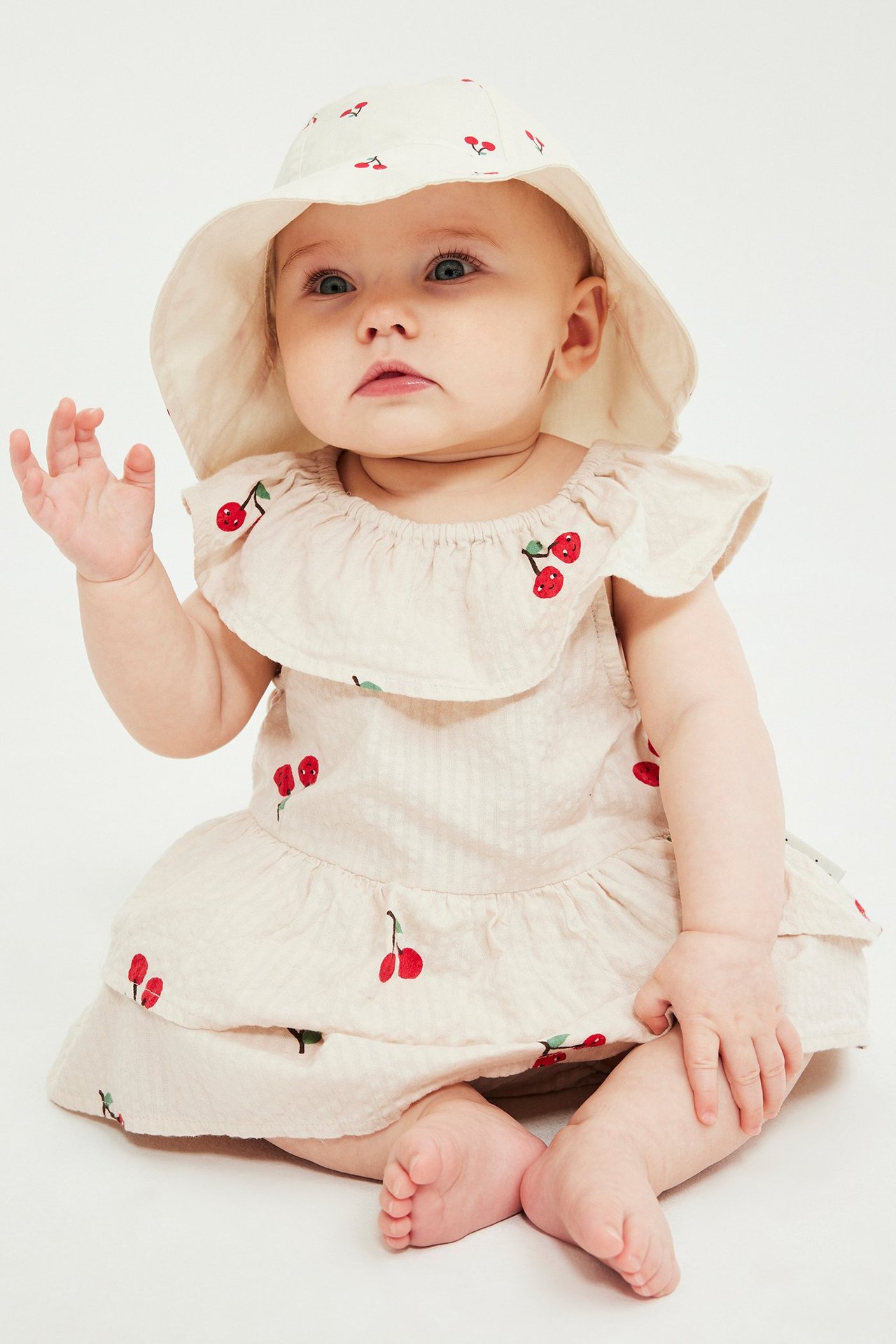 Widok z przodu: niemowlę w beżowej tkanej sukience w wiśnie z falbankami i pasującym kapeluszu przeciwsłonecznym.