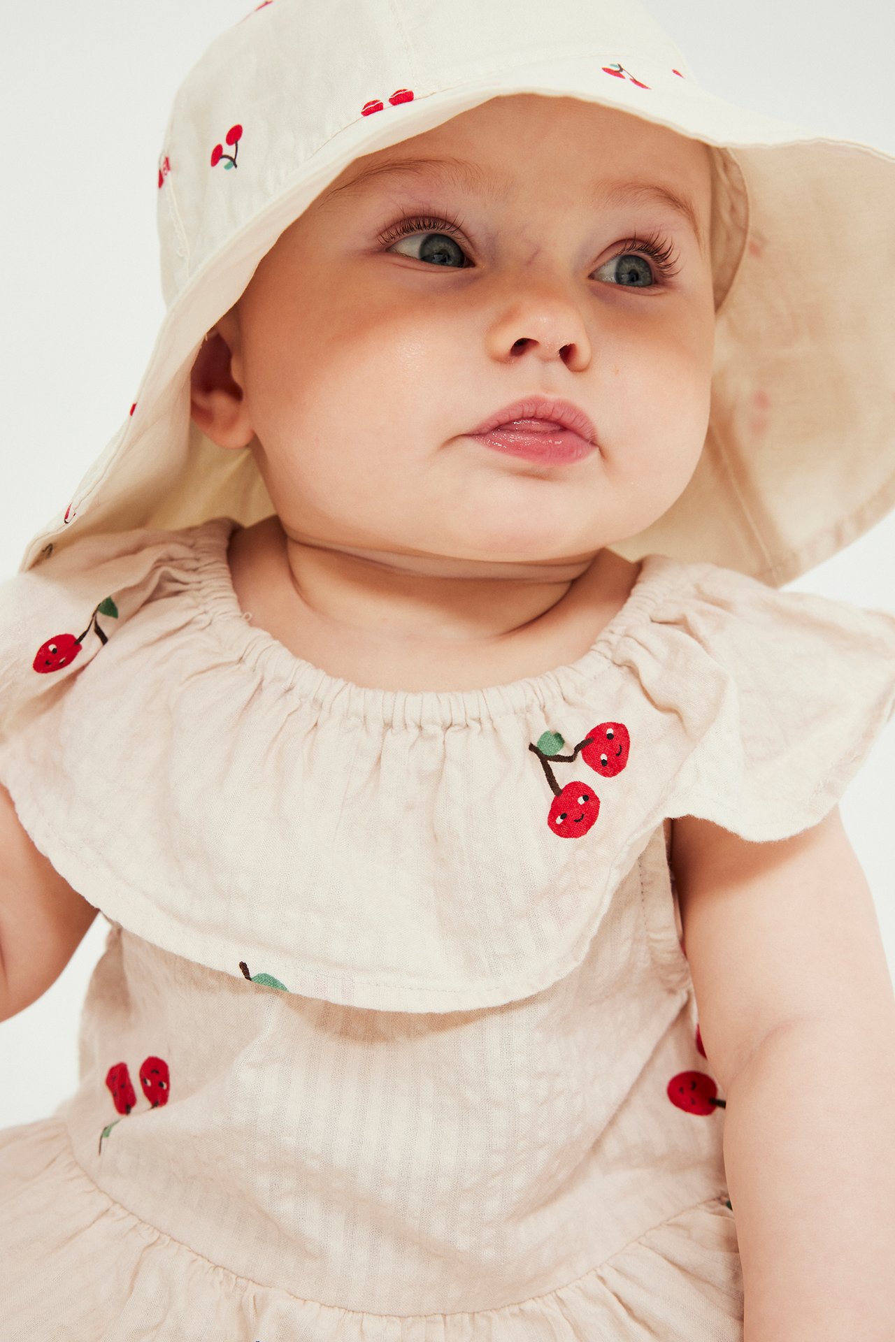 Nærbilde av en baby i en beige, kirsebærmønstret, kortermet seersucker-kjole og matchende hatt.