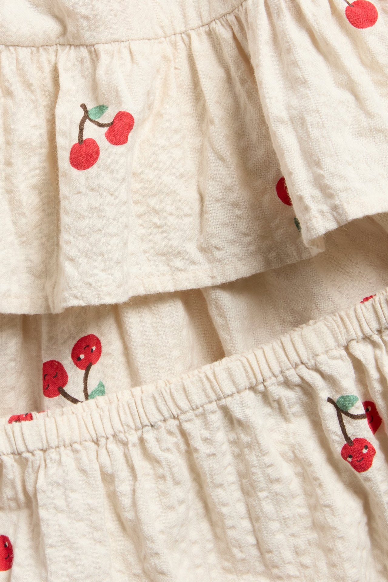Lähikuva beigestä seersucker-vauvanmekosta, jossa kirsikkaprintti ja yhteensopivat joustoresorilliset bloomersit.