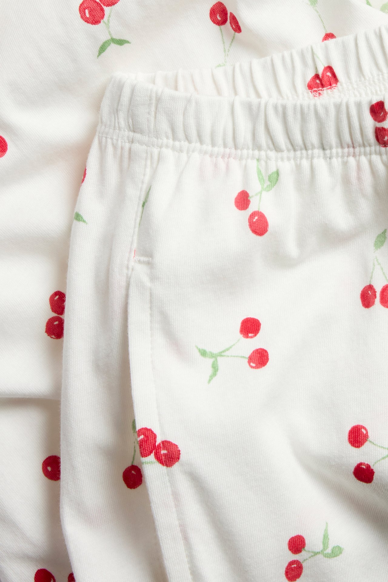 Lähikuva luonnonvalkoisista puuvillaisista pyjamahousuista, joissa punaisia kirsikkaprinttejä, joustava vyötärö ja sivutasku.