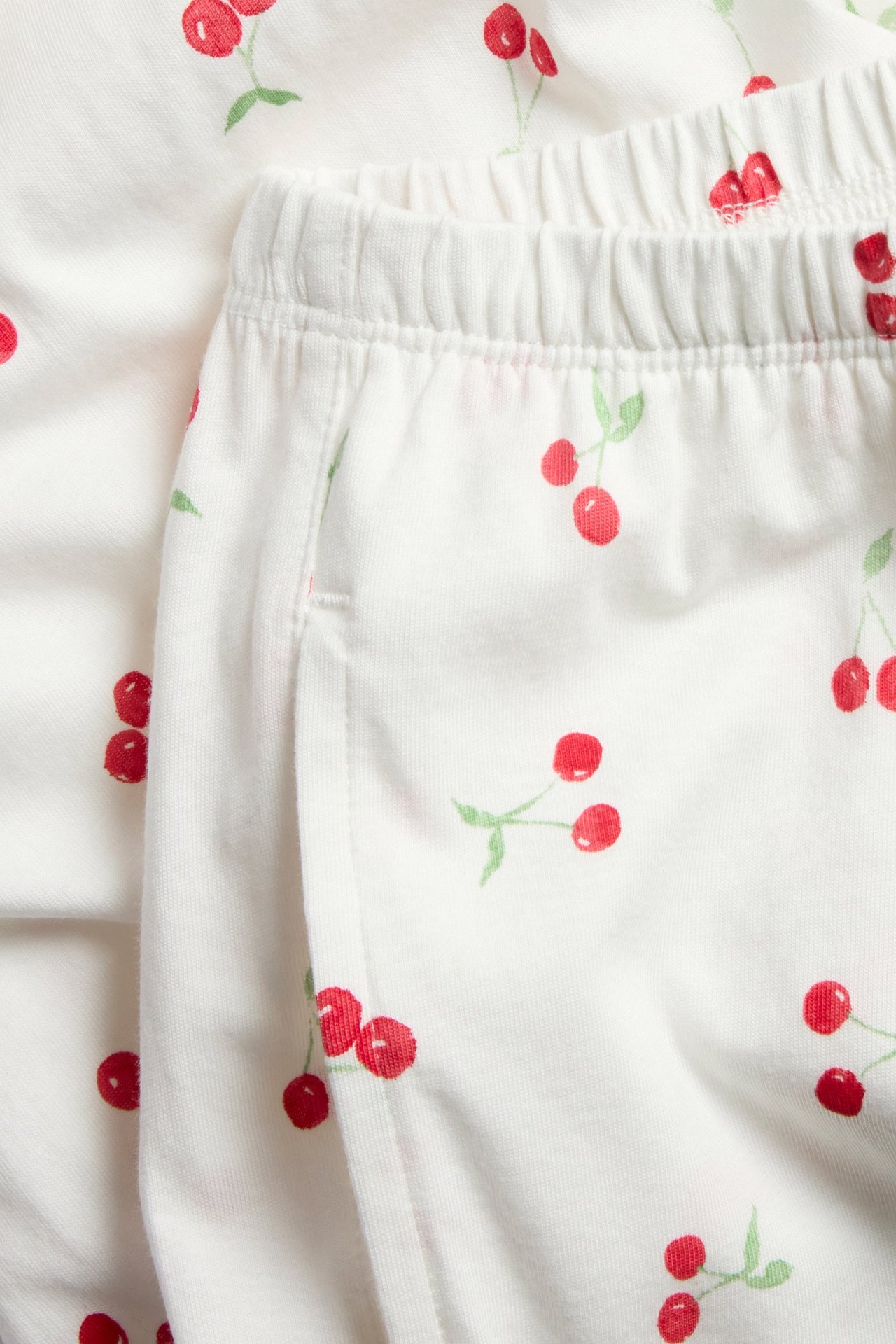 Zbliżenie na off-white bawełniane spodnie piżamowe z czerwonym wzorem w wiśnie, elastyczną talią i kieszenią boczną.