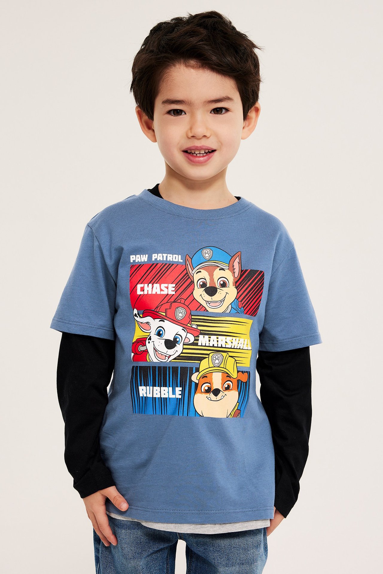 Edestä kuvattu lapsi yllään sininen Paw Patrol puuvilla T-paita mustan pitkähihaisen ja sinisten farkkujen päällä.