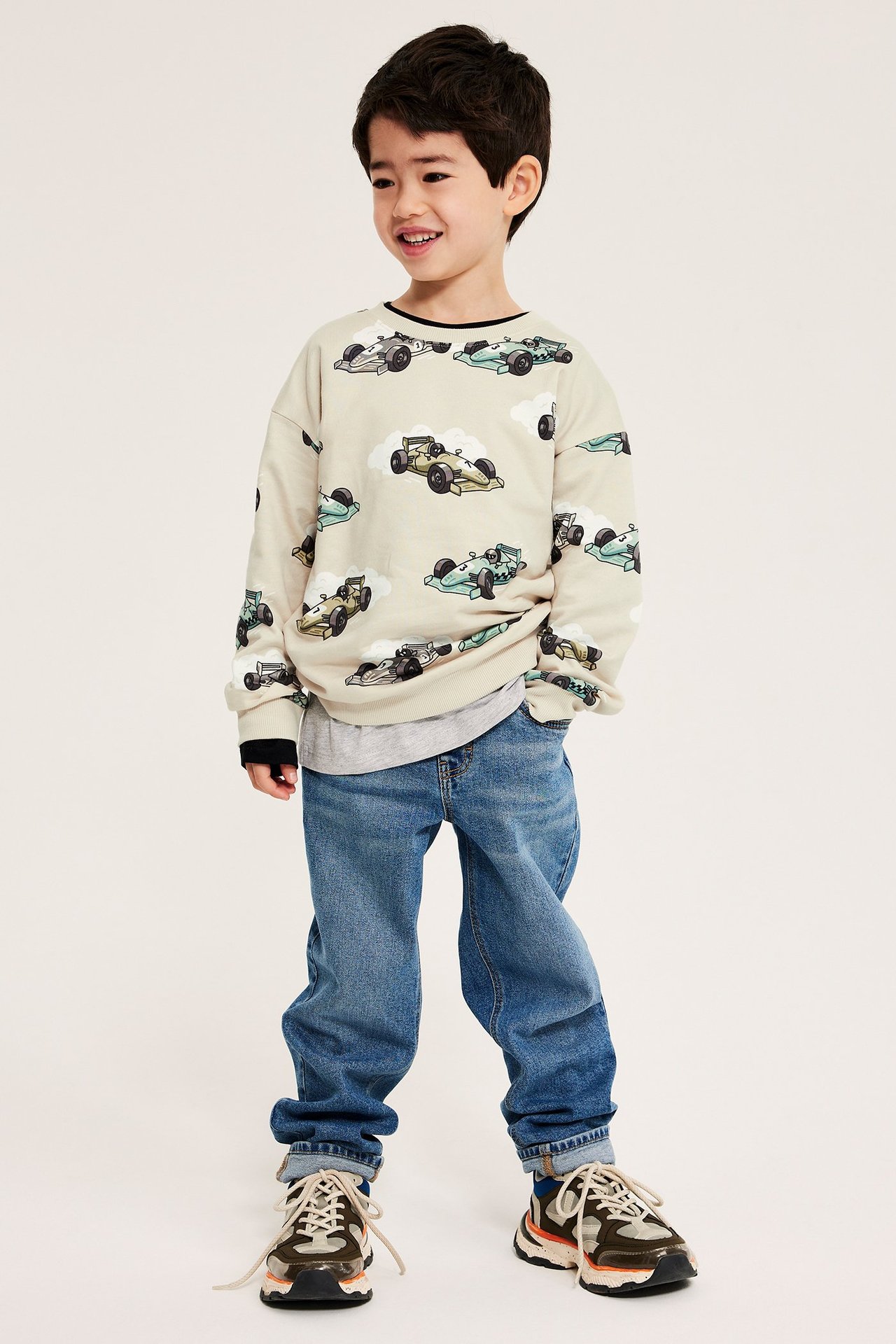 Framifrån: ett barn i beige sweatshirt med biltryck, grå t-shirt, blå jeans och sneakers.