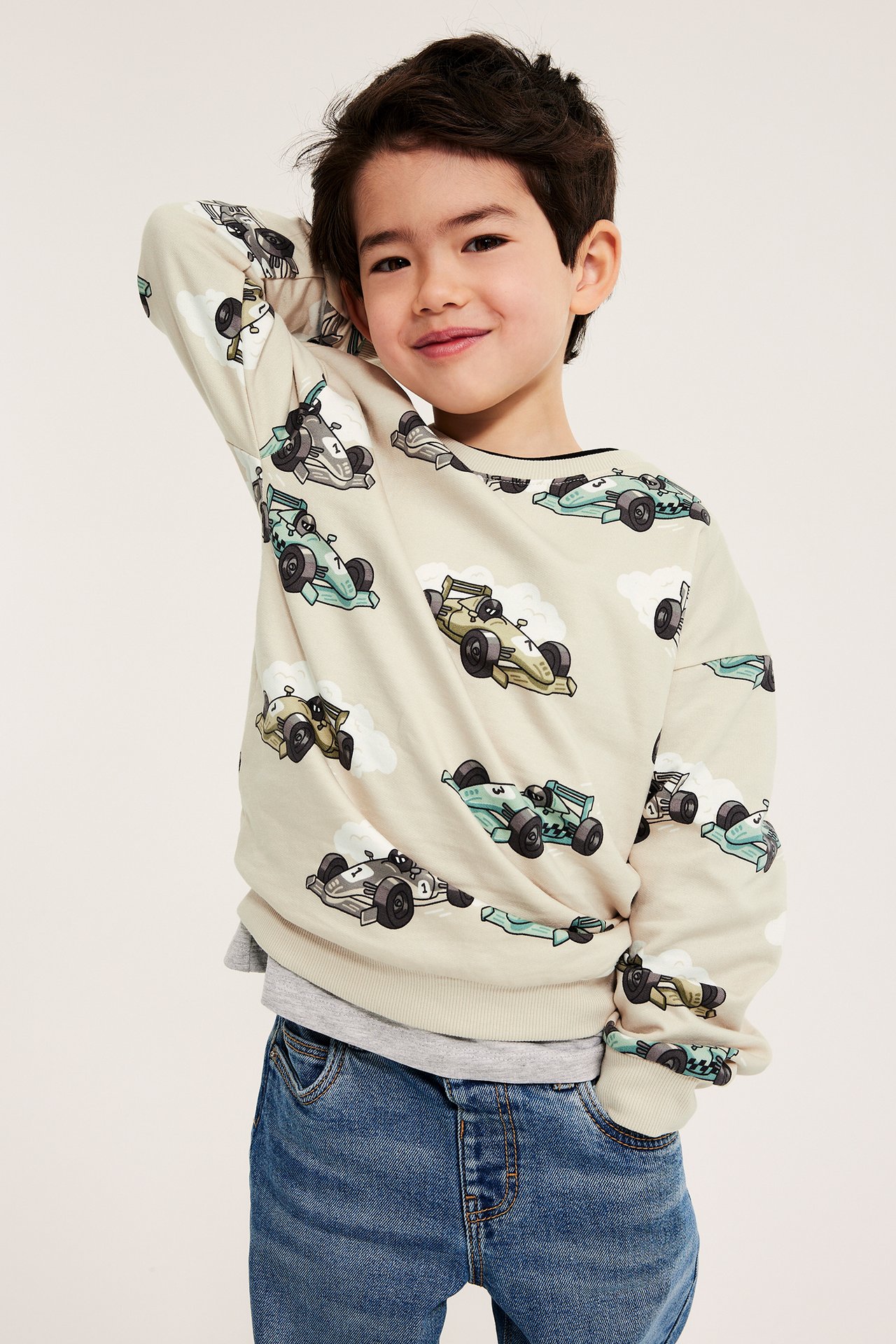 Framifrån: Barn i beige bomulls-sweatshirt med bilmönster, grå t-shirt och blå jeans.