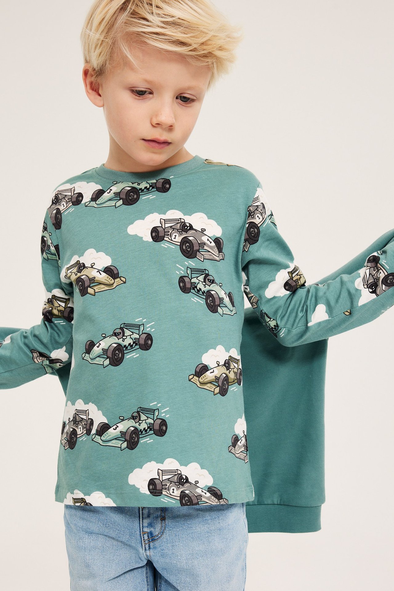Framifrån: Pojke i en ljusgrön långärmad bomulls-t-shirt med bilmönster, med ljusblå jeans.