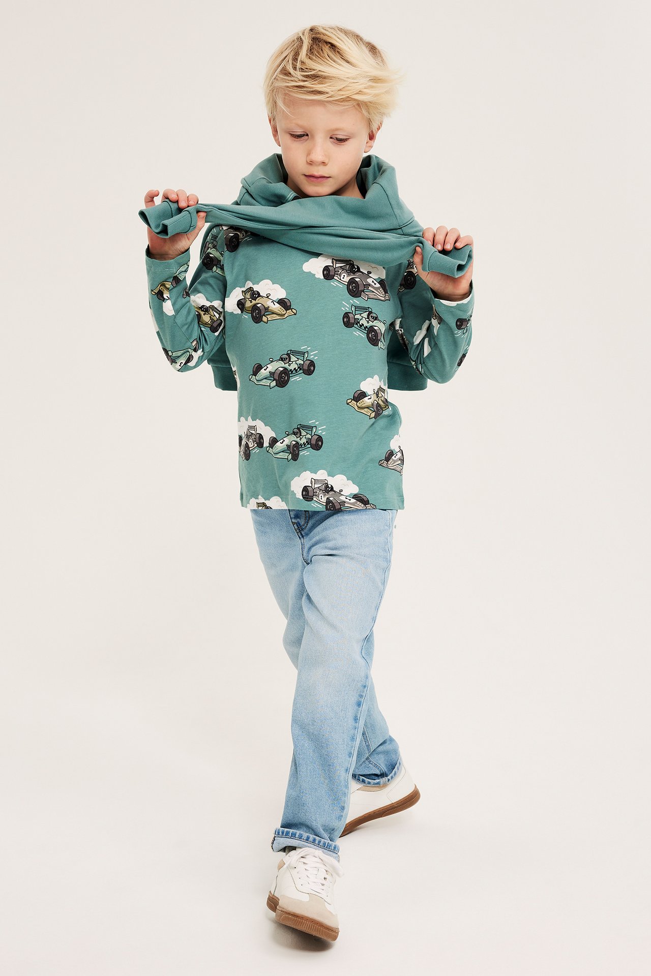 Forfra: et barn drar i en lysegrønn langermet t-skjorte med biltrykk, iført lyseblå jeans og joggesko.