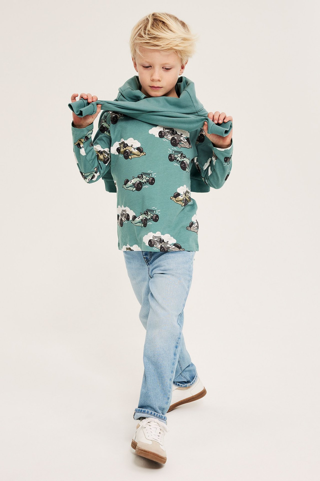 Framifrån: ett barn drar i en ljusgrön långärmad t-shirt med biltryck, bär ljusblå jeans och sneakers.