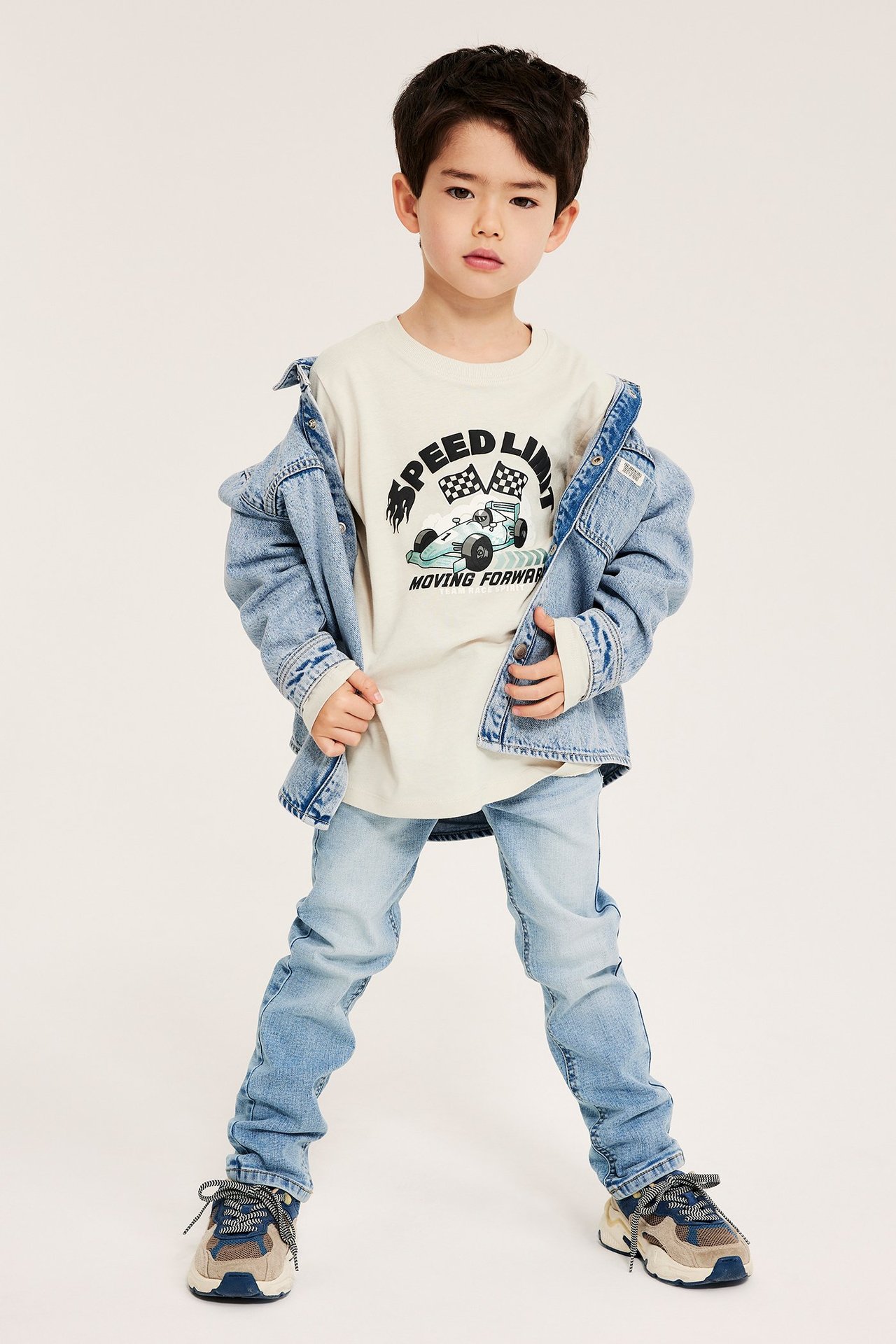 Framifrån: Pojke i beige långärmad t-shirt med biltryck, ljusblå jeansjacka och jeans.