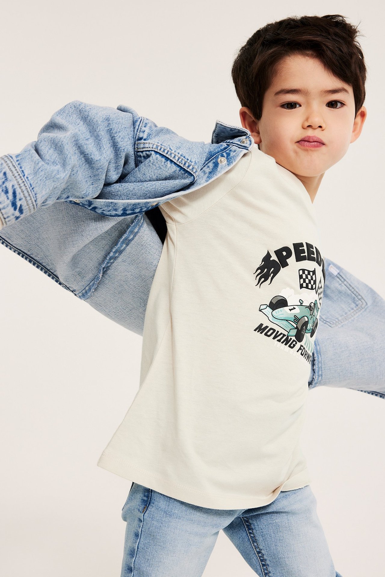 Frontbilde av en gutt i en beige langermet t-skjorte med biltrykk, lysblå jeans og en dongerijakke.