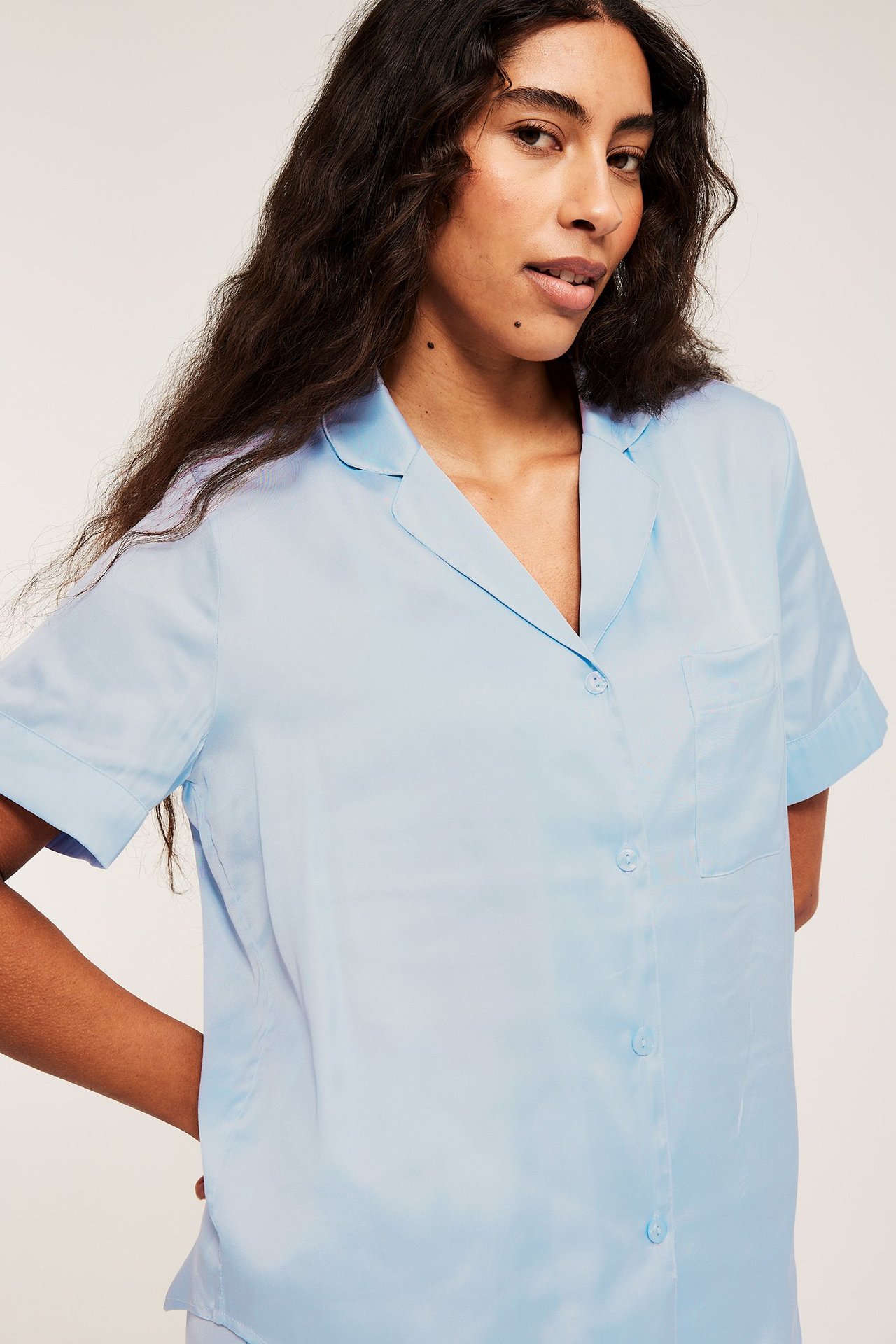 Nærbilde forfra av en kvinne i lys blå viskosesatin pyjamatopp med korte ermer, slagkrage og brystlomme.