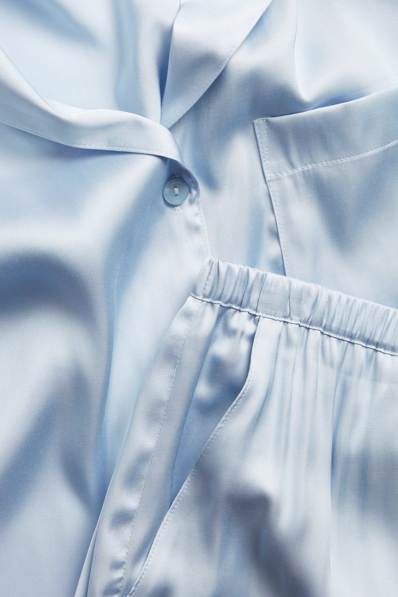 Nærbilde: lyseblå pysjamas i viskosesateng. Toppen har knapp, brystlomme; shorts med strikk i midjen, sidelommer.