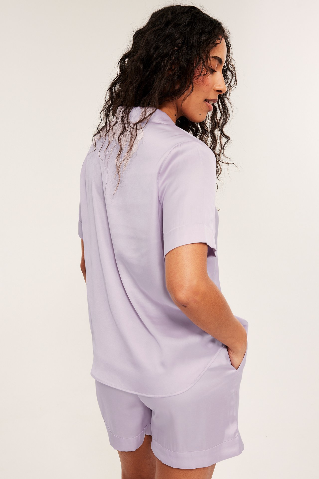 Side-bakfra av kvinne i lys lilla viskose-satin pyjamatopp og shorts, med hånden i lommen.