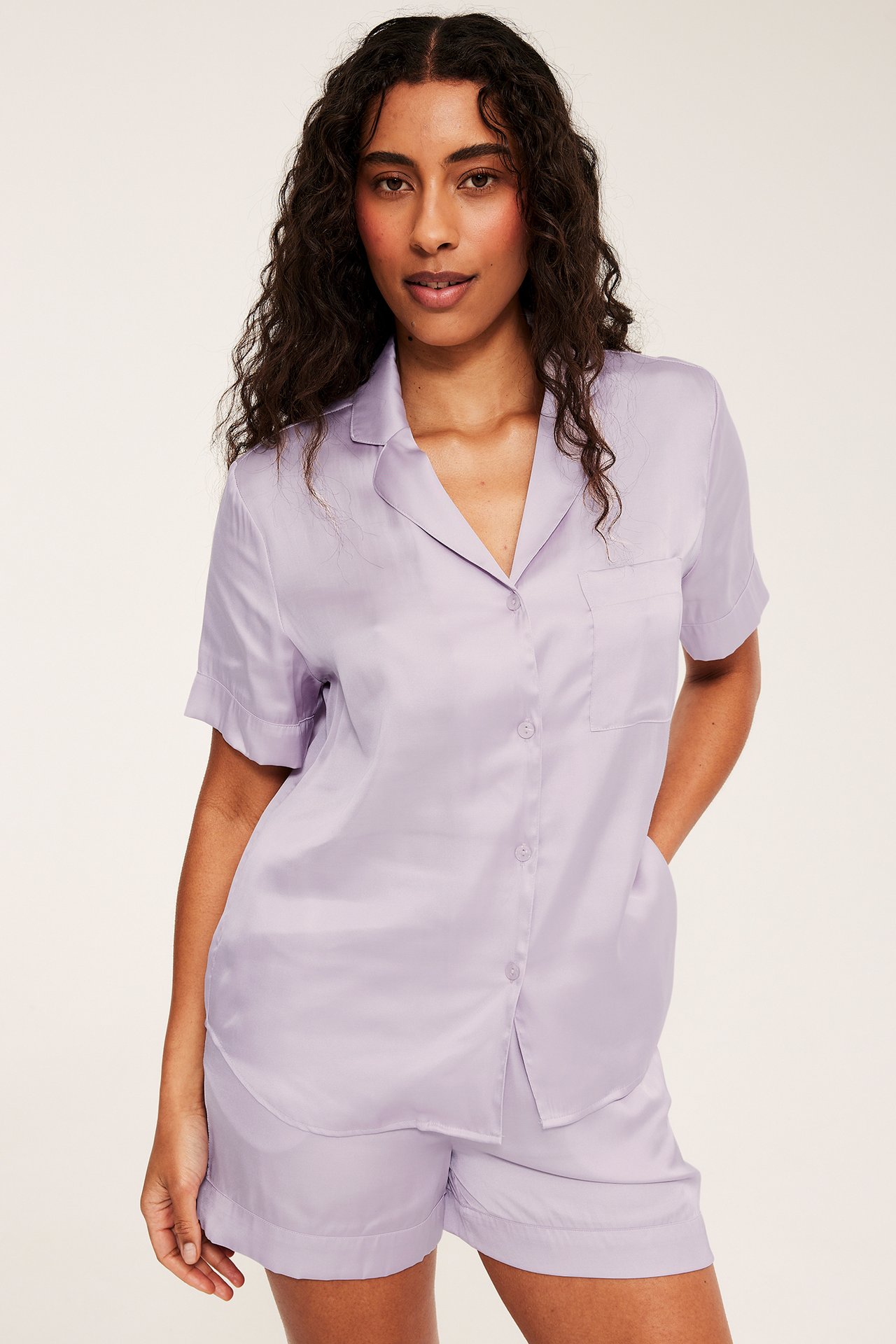 Edestä: nainen vaalealilassa viskoosimix-pyjamasissa, napillinen paita taskulla ja yhteensopivat shortsit.