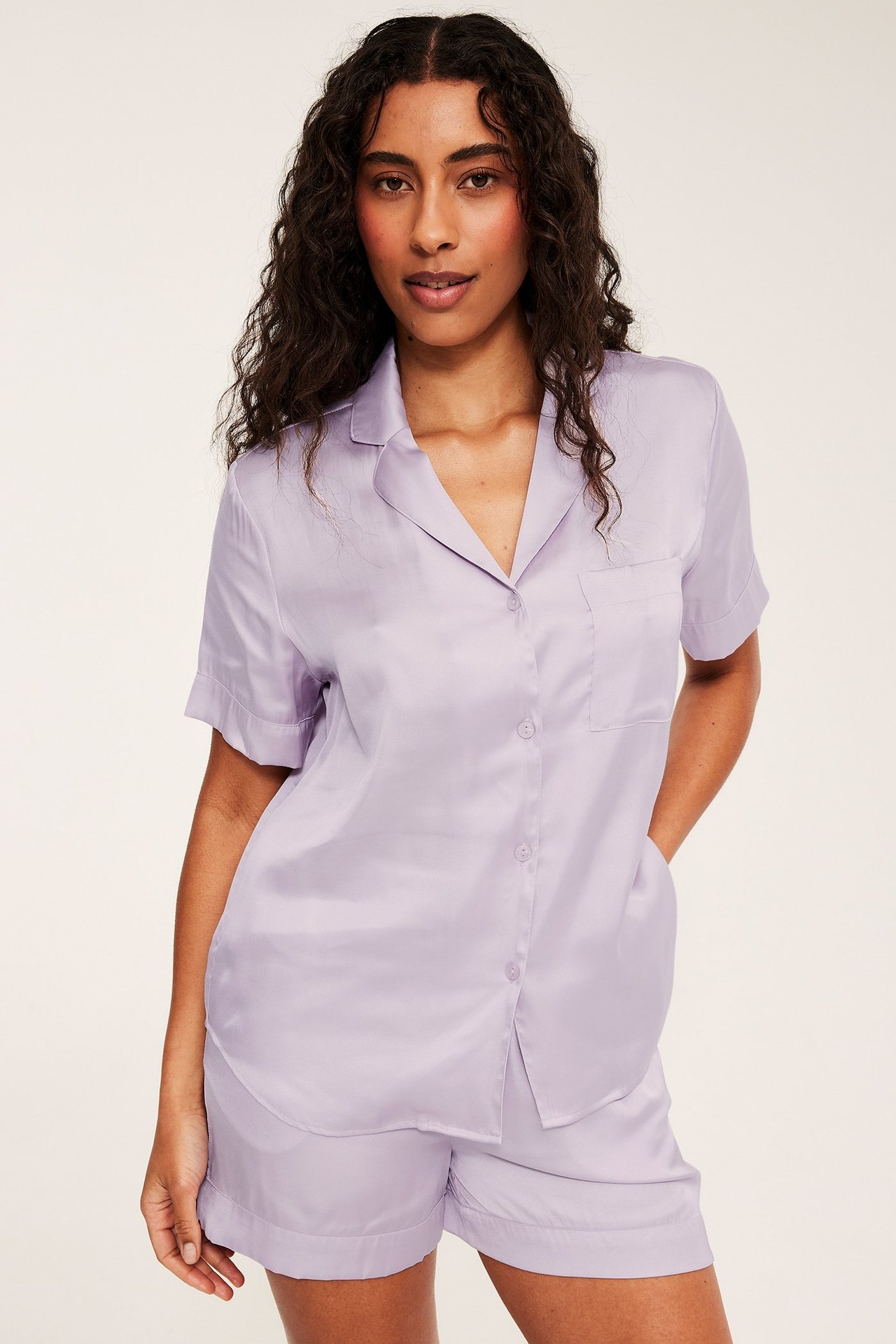 Widok z przodu kobiety w jasnoliliowym piżamowym zestawie z wiskozy: koszula z guzikami i kieszenią oraz szorty.