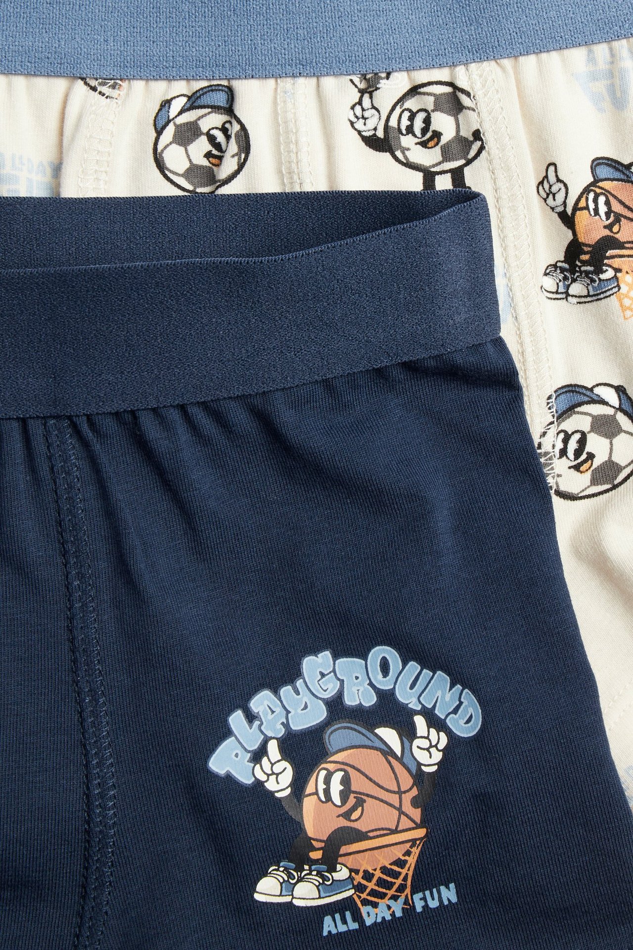 Lähikuva kahdesta parista lasten boksereita: tummansiniset koripallopainatuksella, luonnonvalkoiset jalkapallokuviolla.