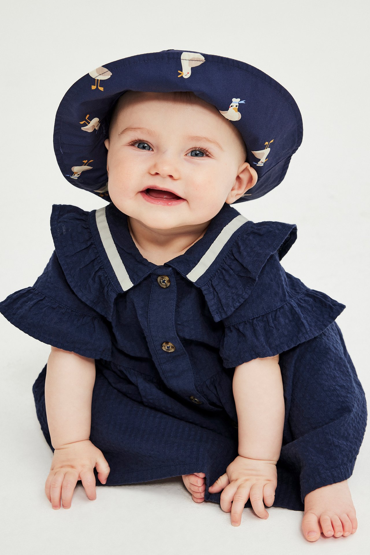 Forfra: En baby i en marineblå seersucker-kjole med volanger, knapper og en mønstret solhatt.