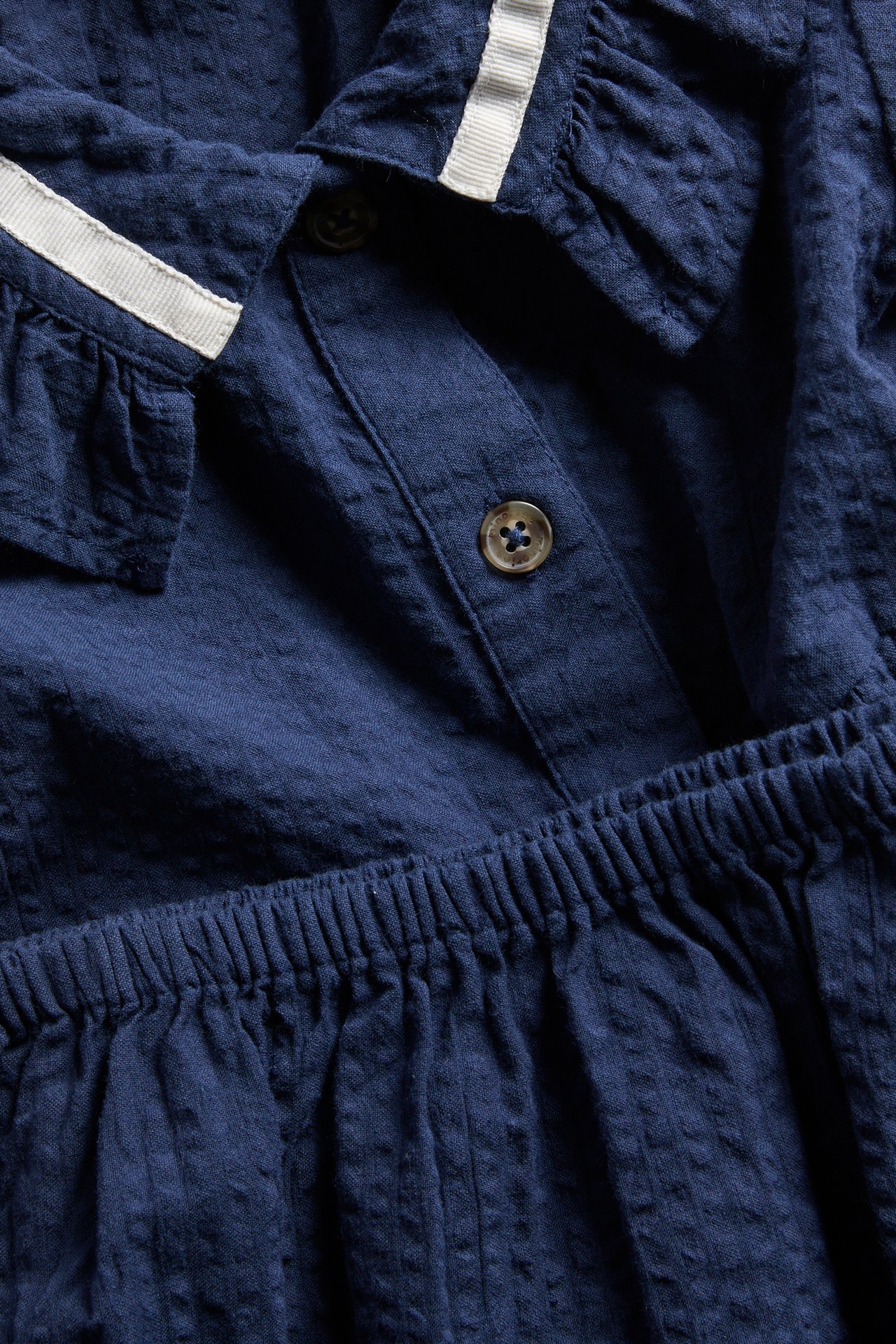 Nærbilde forfra: mørkeblå babykjole i seersucker med volangkrage, hvit kant og knapper foran.
