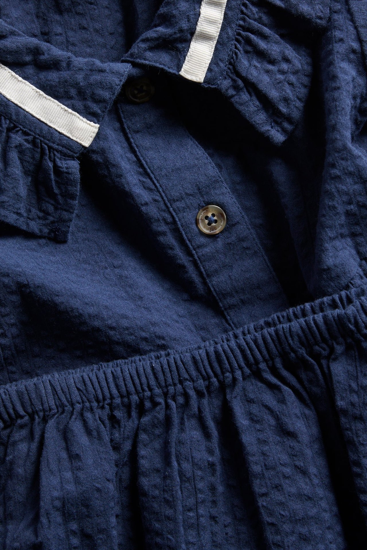 Zbliżenie: granatowa sukienka niemowlęca z seersuckera z falbankowym kołnierzem, białym wykończeniem i guzikami.