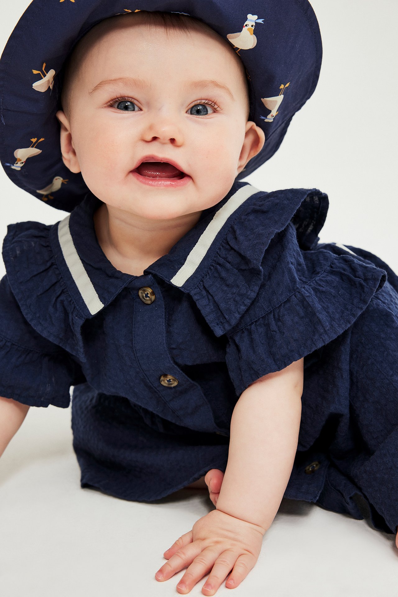 Nærbilde av en baby i blå seersucker-kjole med volanger og marineblå hatt med fugletrykk.