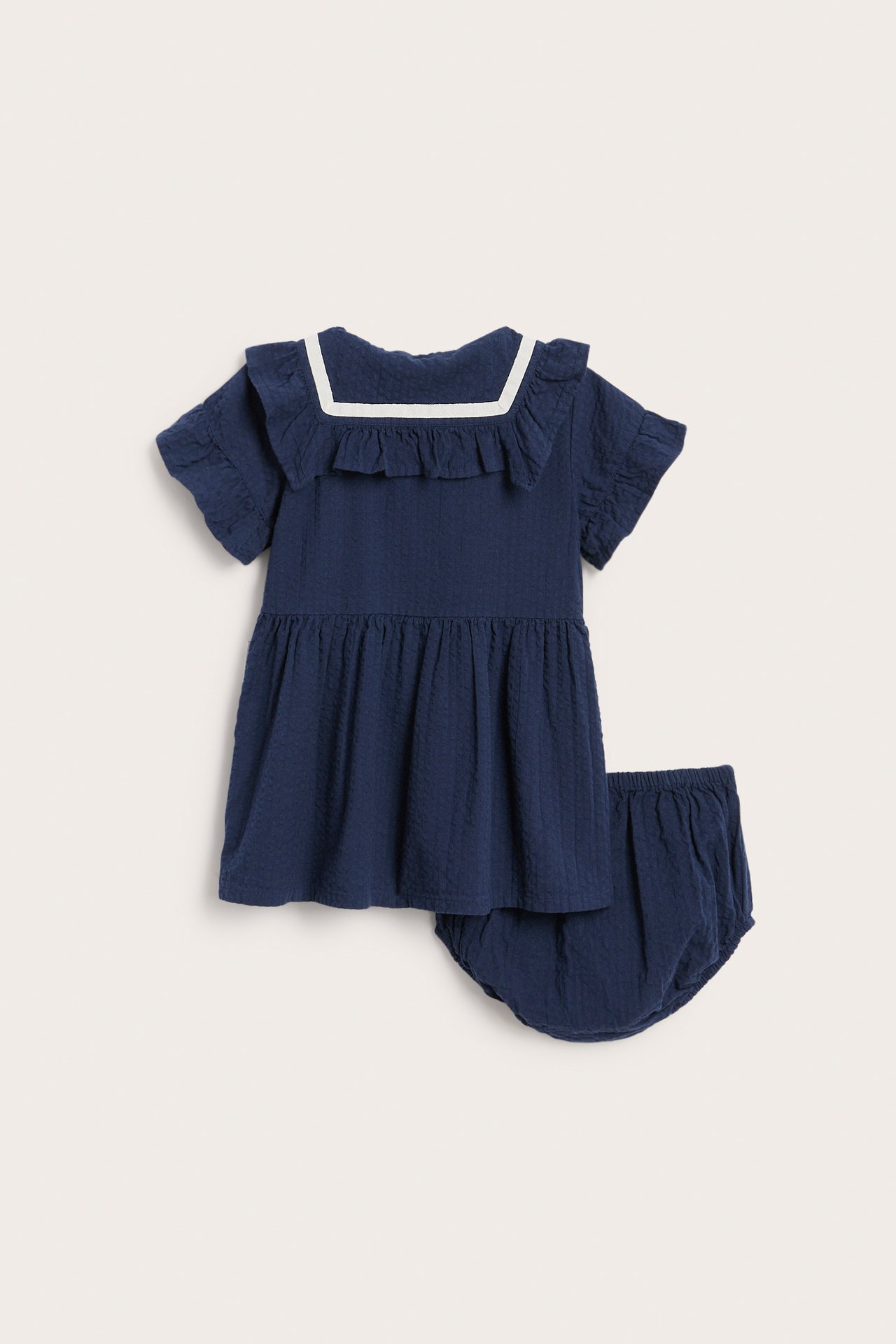 Framifrån: Mörkblå texturerad babyklänning med vitkantad fyrkantig krage och matchande bloomers.