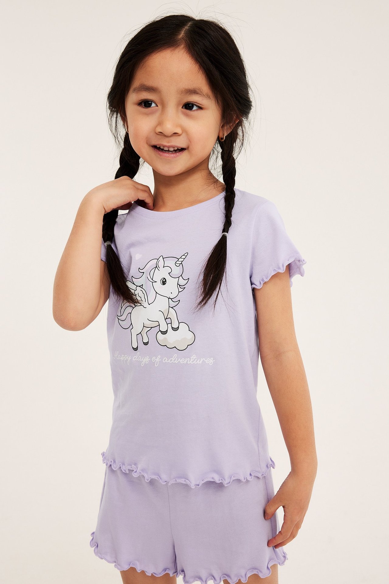 Edestä: Lapsi vaaleanlilassa puuvillapyjamasetissä, yksisarvistoppi ja yhteensopivat frillashortsit.