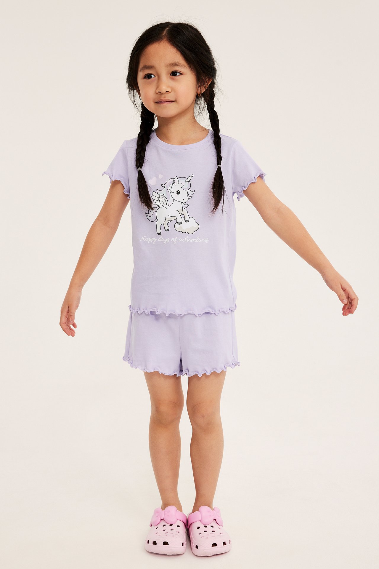 Frontbilde: Et barn har på seg et lyslilla bomullspyjamassett med enhjørning-t-skjorte, rysjeshorts og rosa tresko.
