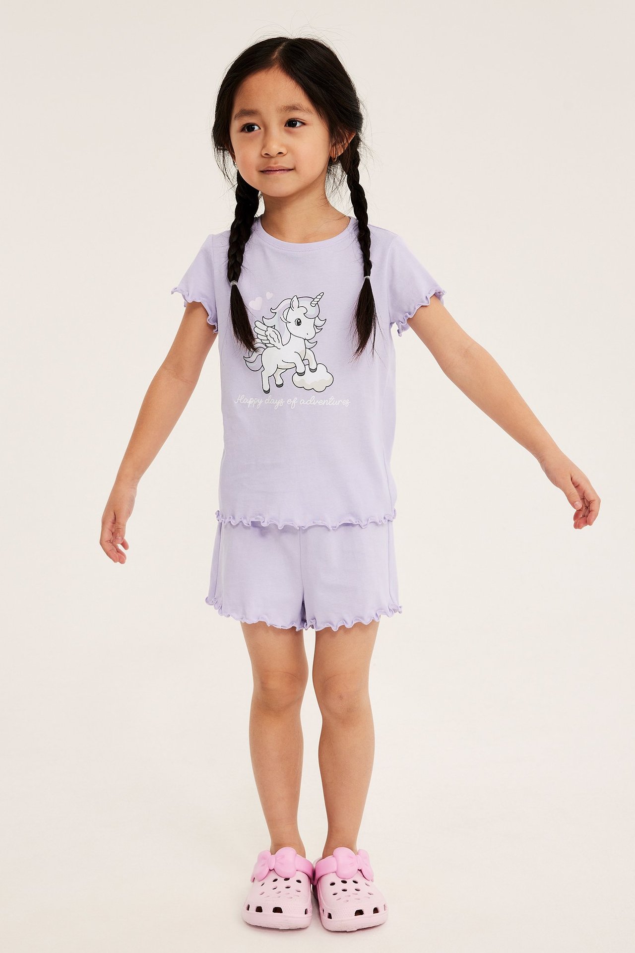 Edestäpäin: Lapsi yllään vaaleanliila puuvillapyjamasetti, jossa yksisarvispaita, röyhelöshortsit ja vaaleanpunaiset pistokkaat.