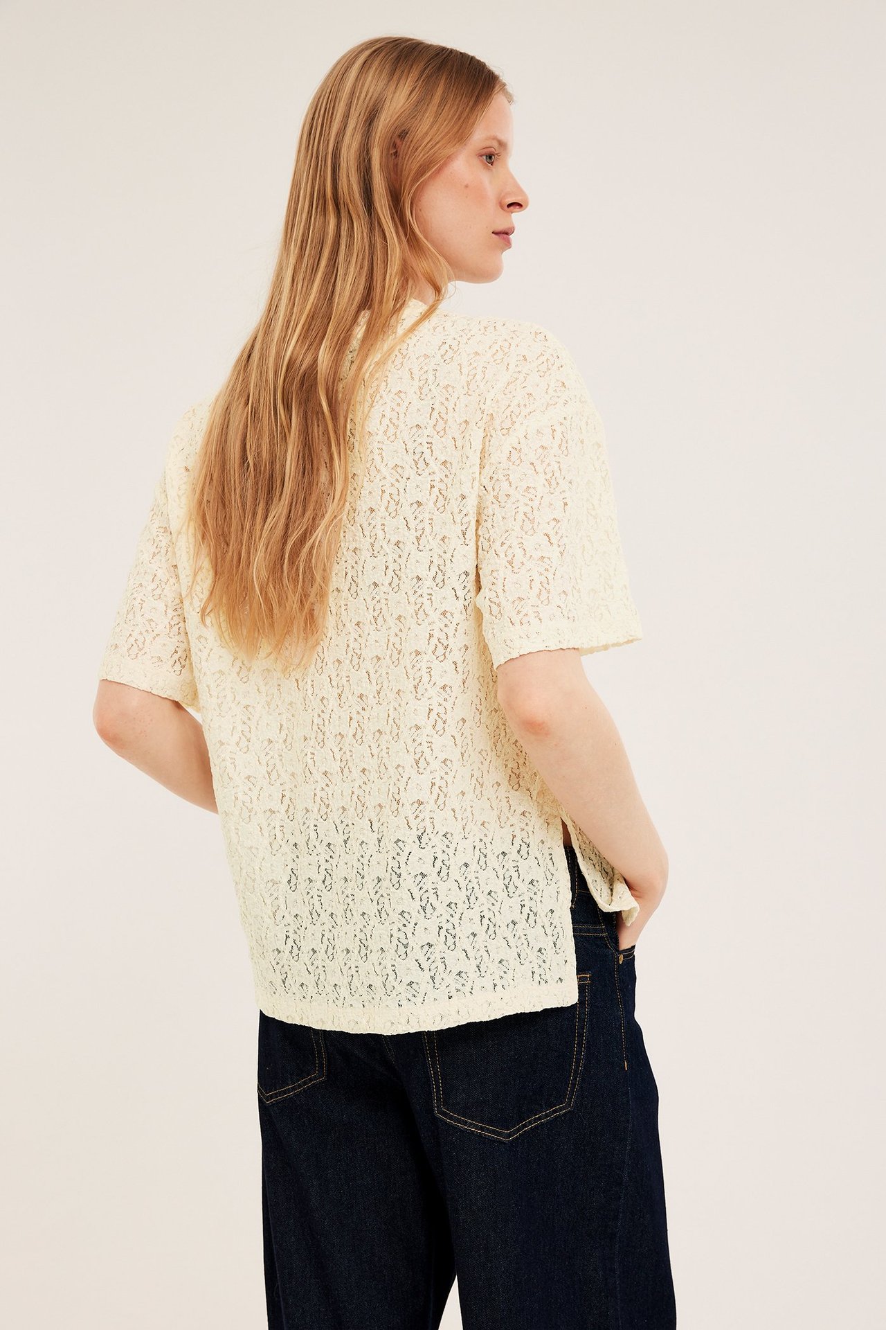 Nainen takaapäin yllään luonnonvalkoinen, väljä, lyhythihainen pitsitoppi ja tummansiniset farkut.