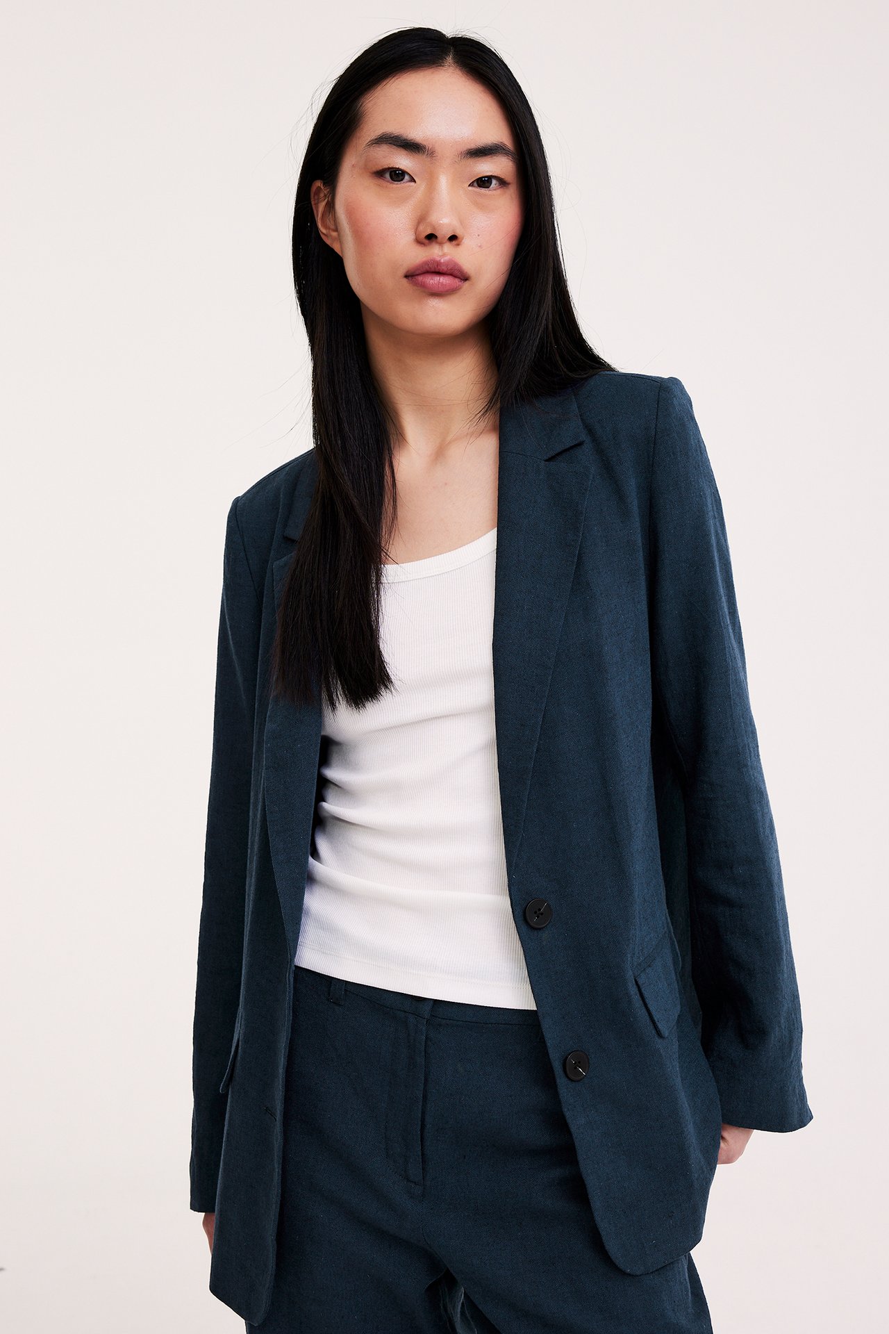 Widok z przodu kobiety w otwartej, granatowej melanżowej marynarce lnianej, białym prążkowanym topie i pasujących spodniach.