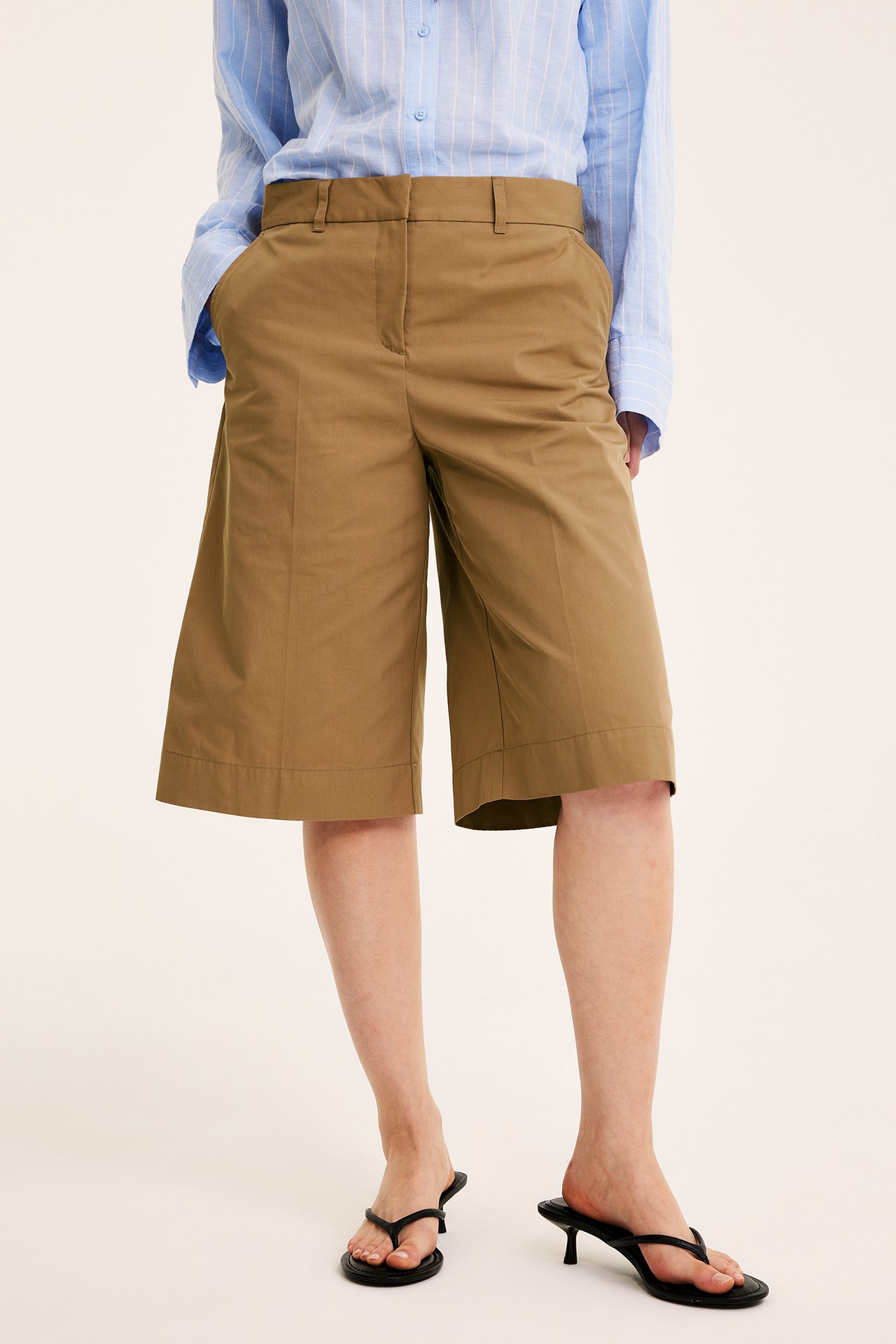 Frontbilde av kvinne i vide beige bermudashorts, lys blåstripete skjorte og svarte sandaler med hæl.