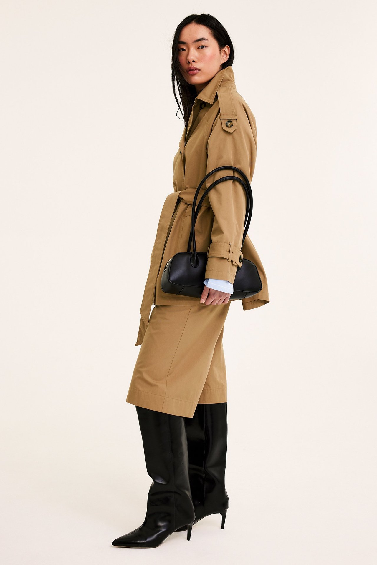 Widok z przodu-boku kobiety w beżowych bermudach do kolan, pasującym trenczu, trzymającej czarną torebkę, czarne kozaki.