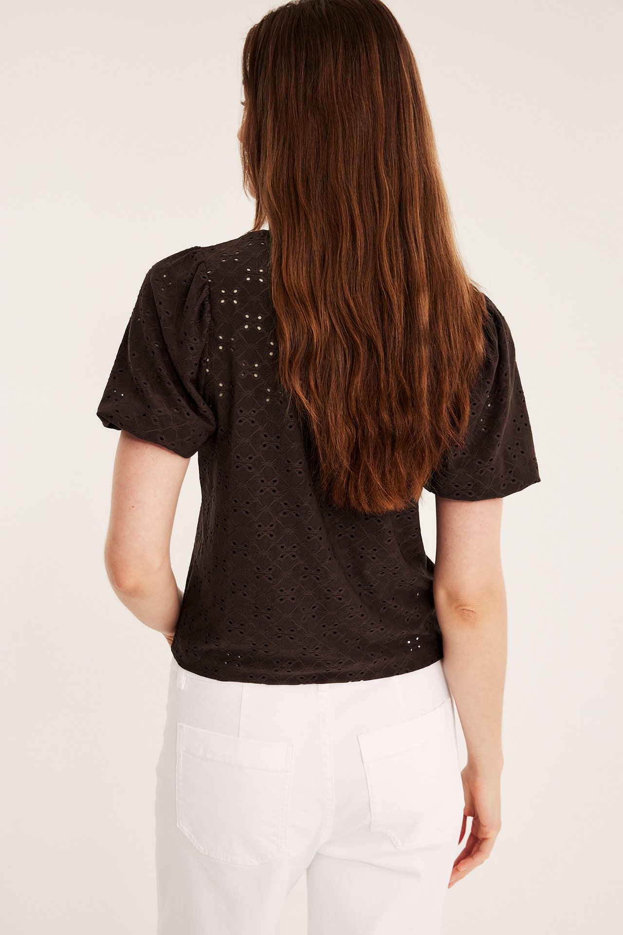 Bakfra: Kvinne i mørk brun broderie anglaise topp med puffermer og hvite bukser.