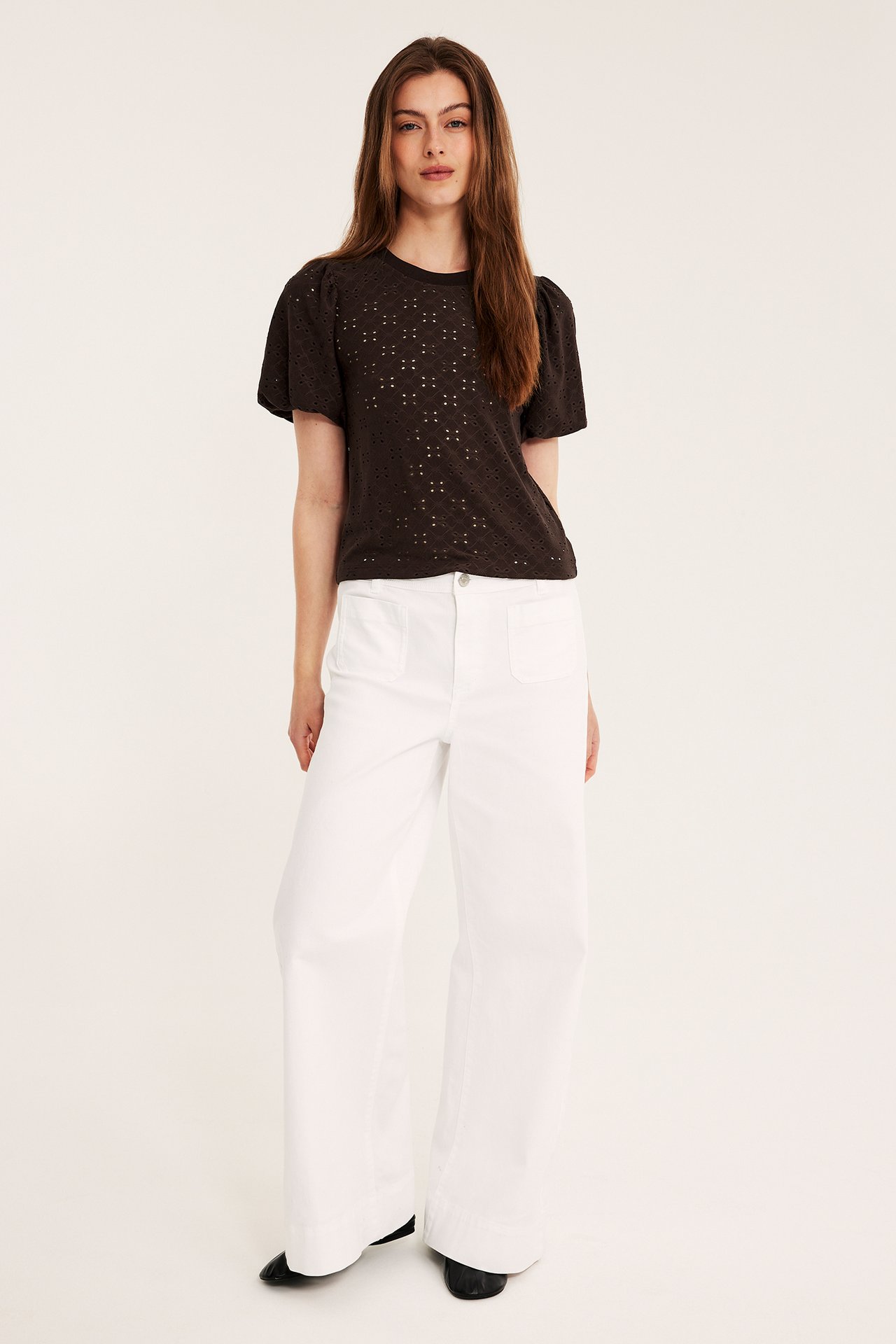 Kvinna bär mörkbrun broderie anglaise topp med puffärm, vita vida byxor och svarta platta skor.