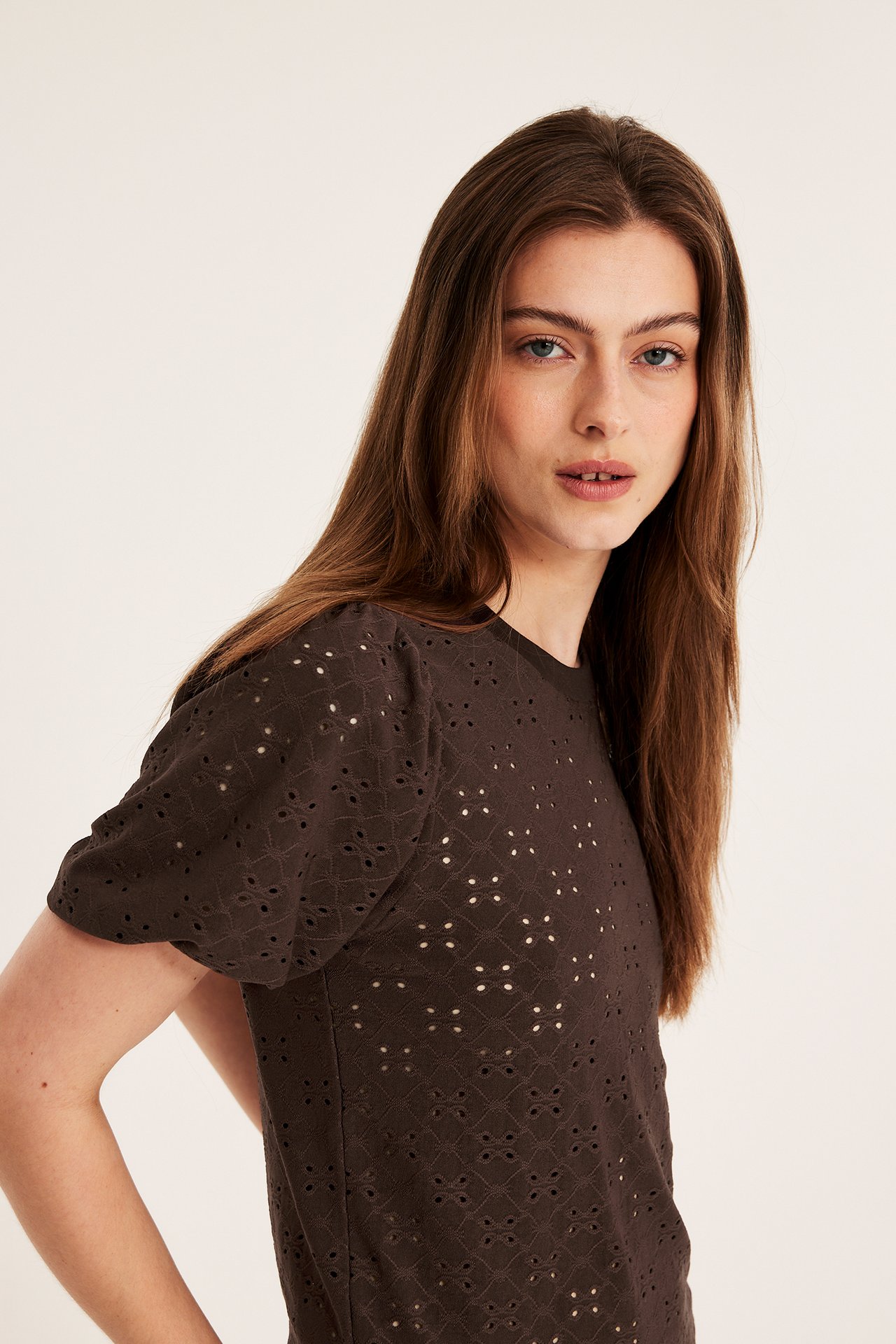 Närbild framifrån av en kvinna som bär en mörkbrun kortärmad topp med puffärmar och broderie anglaise mönster.