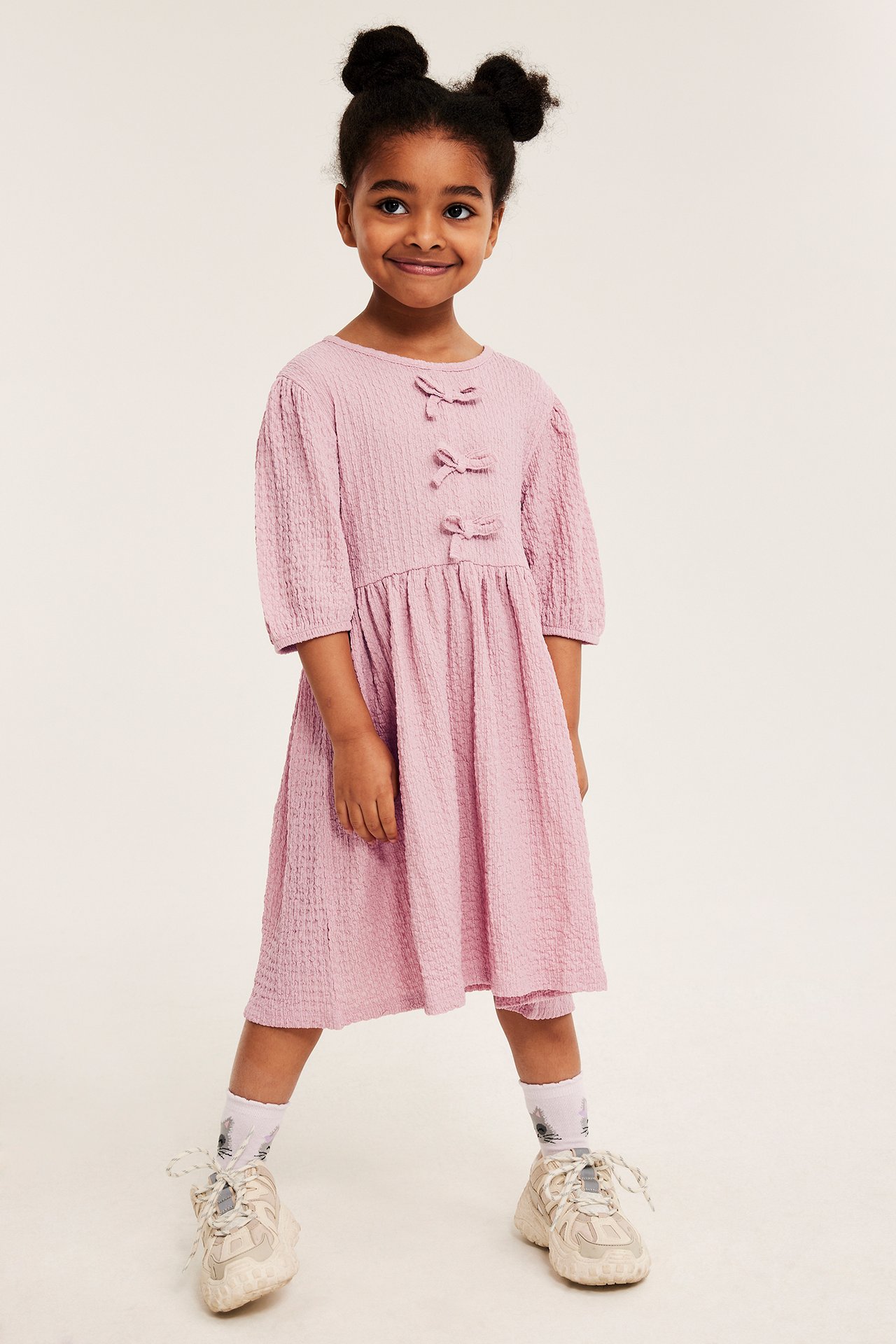 Frontutsikt: barn i lyserosa krinklet kjole med sløyfer, hvite mønstrede sokker og klumpete beige joggesko.