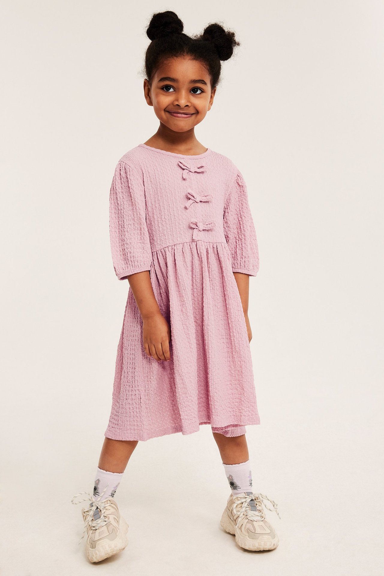 Edestäpäin: lapsi vaaleanpunaisessa rypytetyssä mekossa, jossa rusetit, valkoiset kuviolliset sukat ja paksupohjaiset lenkkarit.