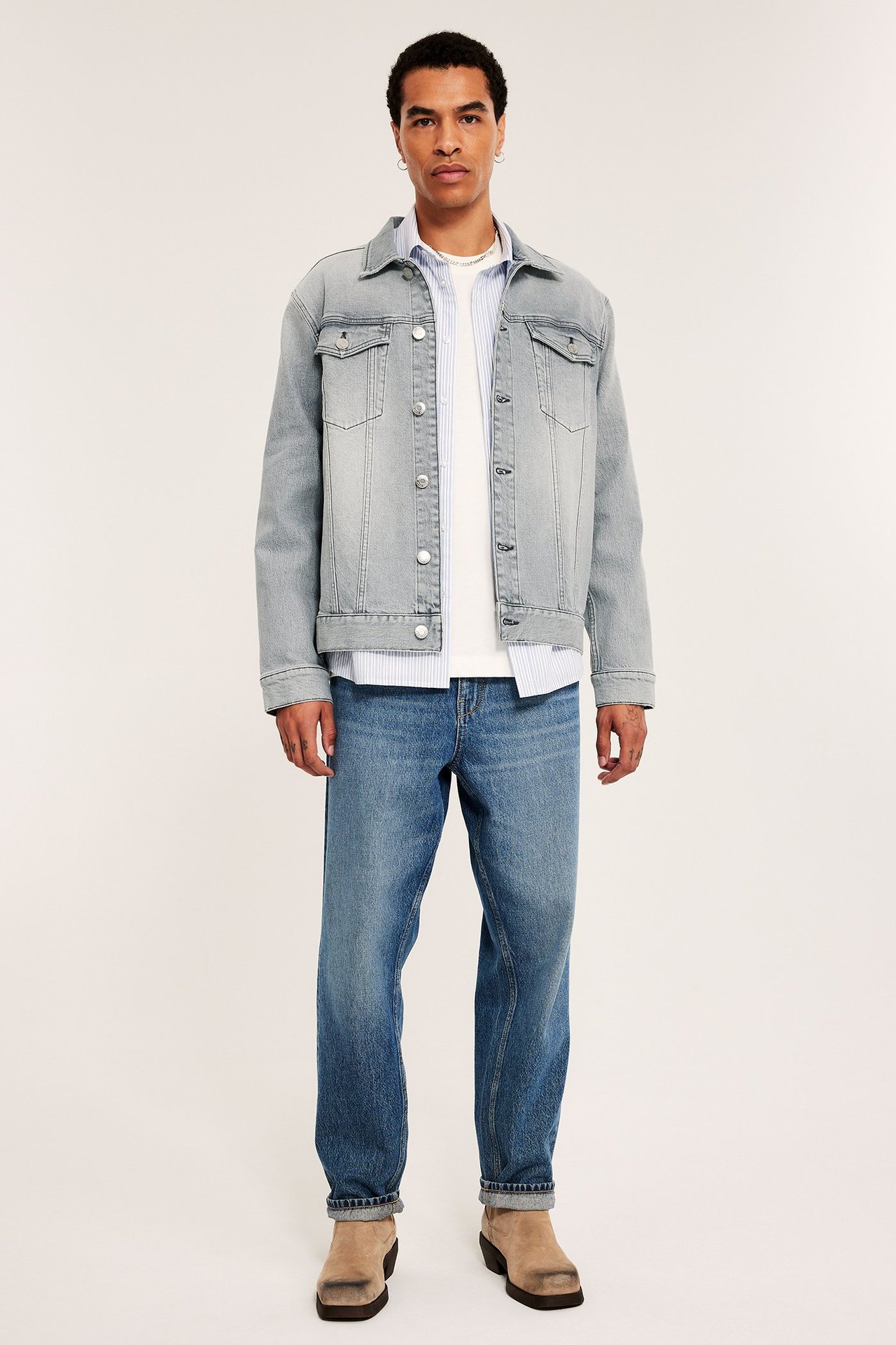 Widok z przodu: mężczyzna w jasnoszarej kurtce dżinsowej z guzikami, niebieskich dżinsach i beżowych butach.