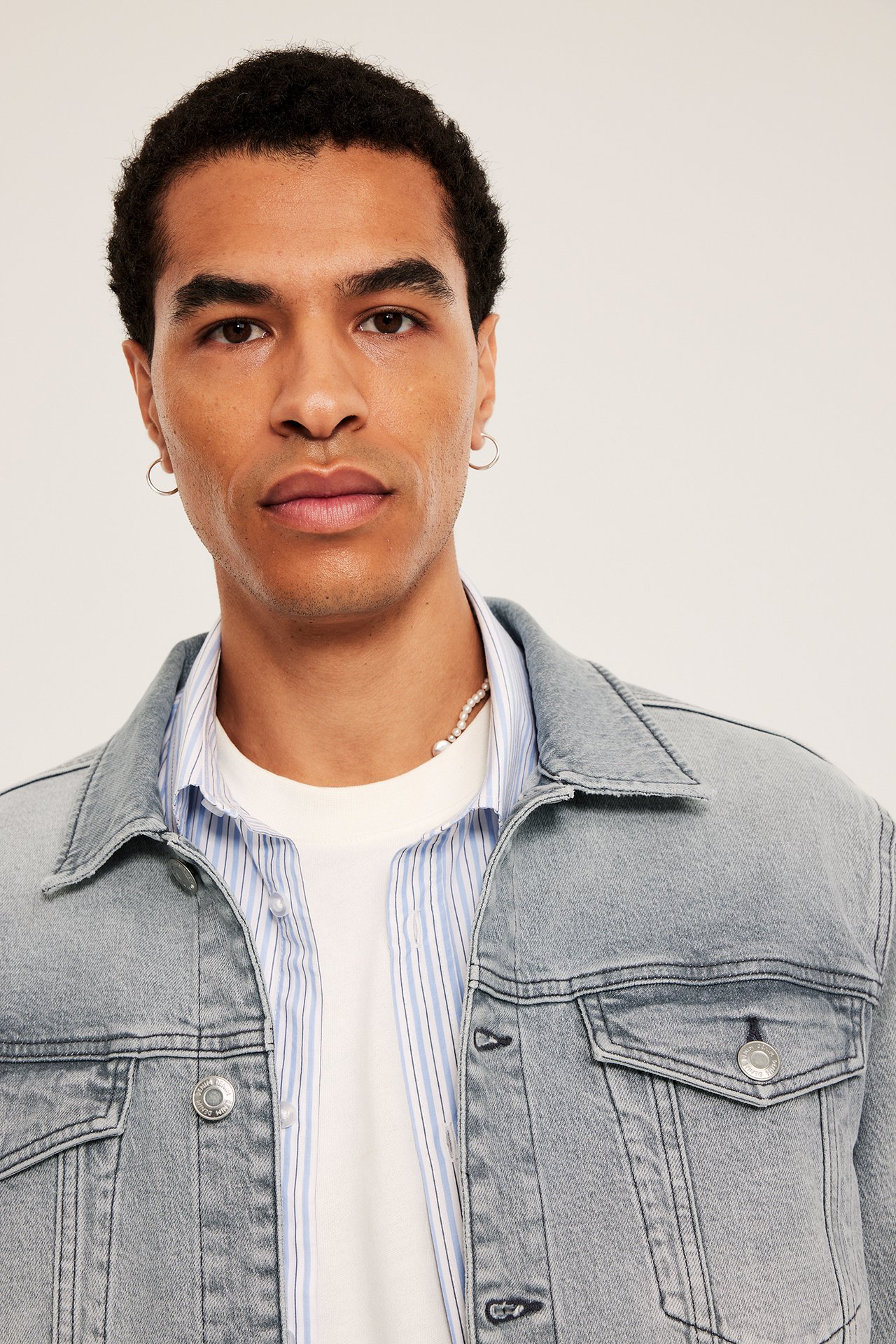 Mann i lys grå dongerijakke med knapper, brystlommer, over stripete skjorte. Perlekjede synlig. Sett forfra.