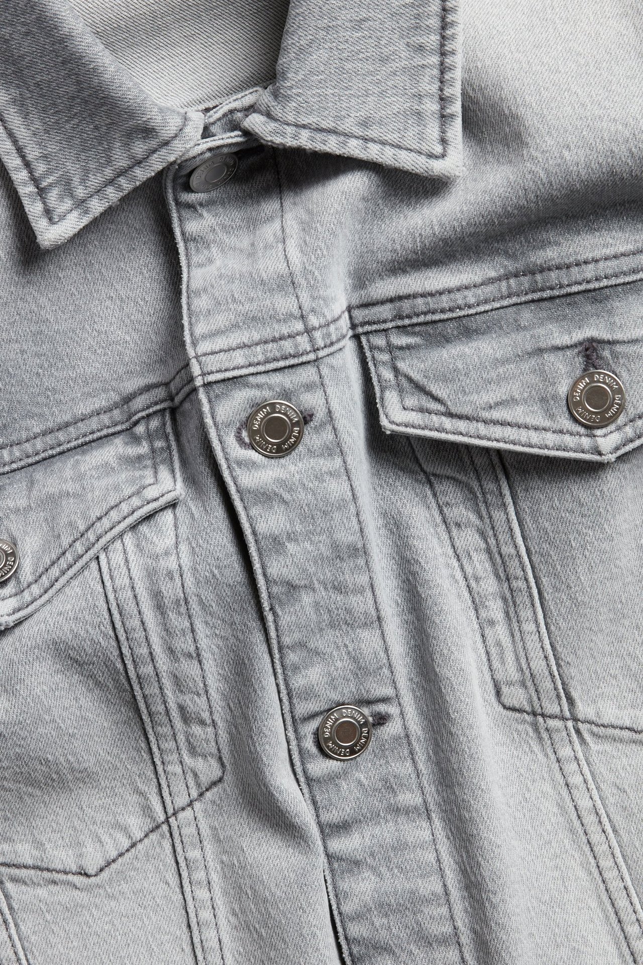 Nærbilde forfra av en lys grå dongerijakke for menn med metallknapper og to brystlommer.