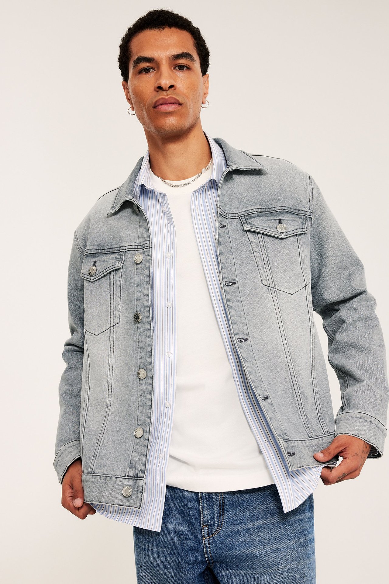 Mężczyzna w jasnoszarej kurtce jeansowej, białym T-shircie, koszuli w paski i niebieskich dżinsach.