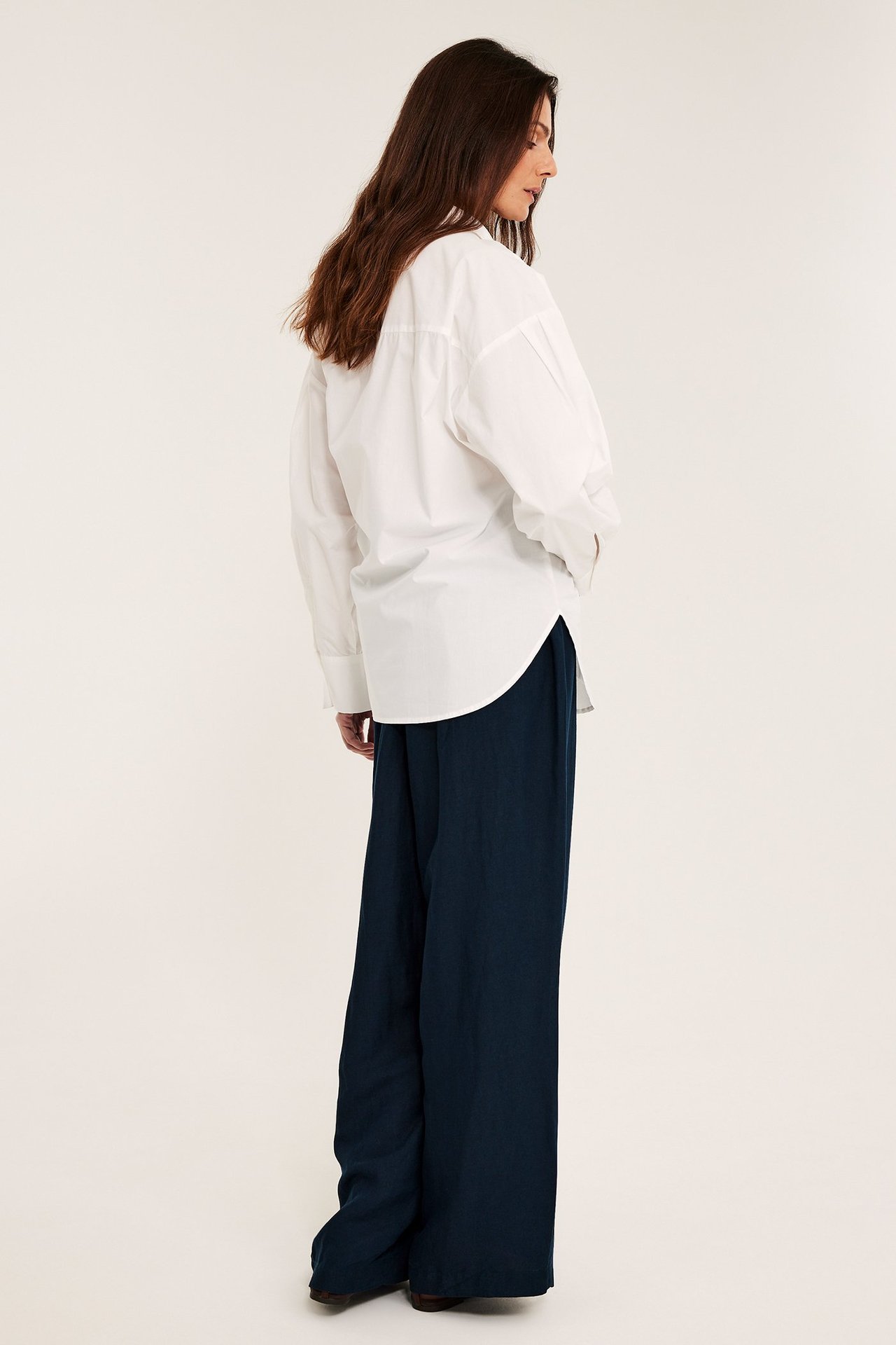 Widok z boku, od tyłu kobiety w ciemnoniebieskich, szerokich spodniach z lnu i białej koszuli oversize.