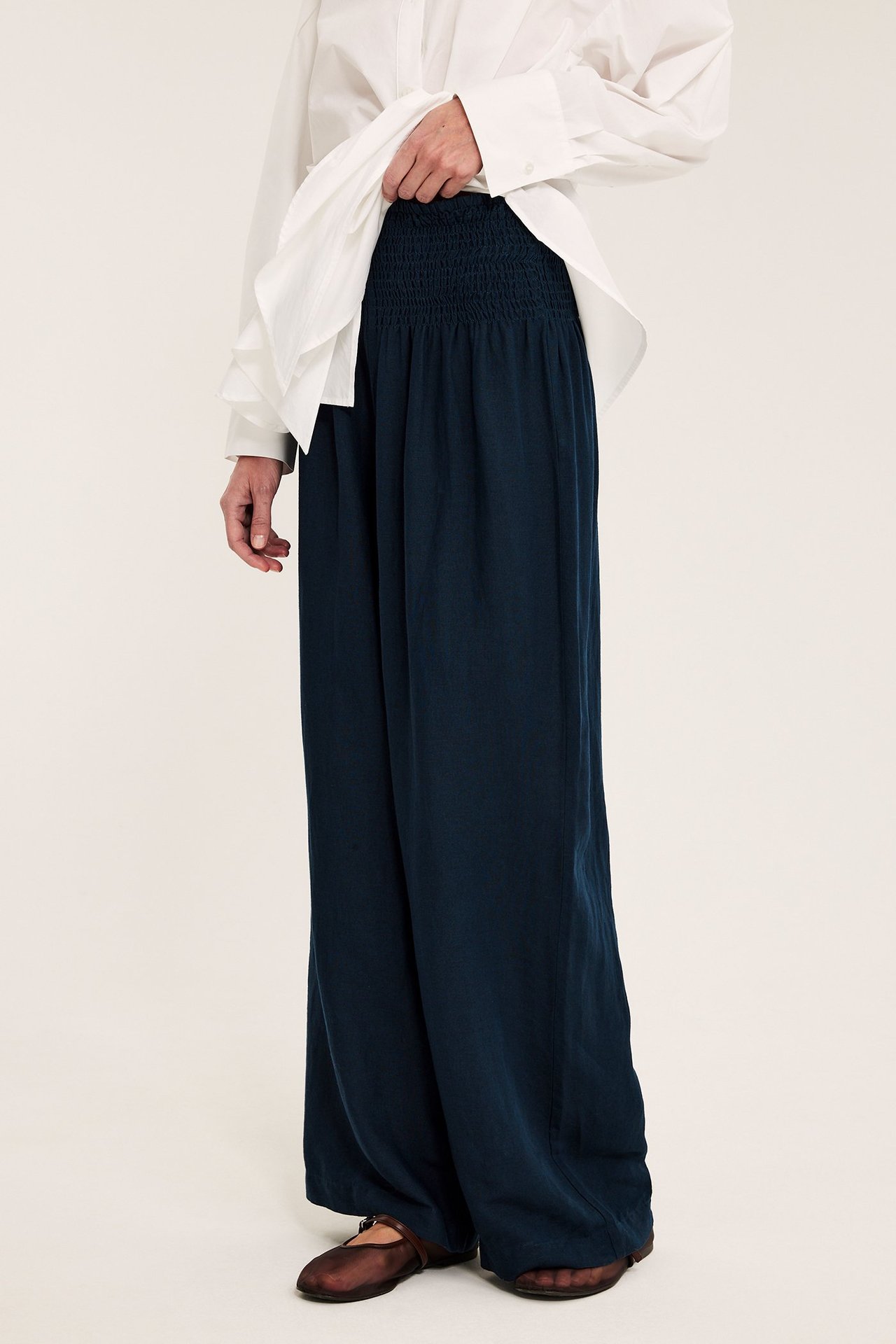 Sidebilde: Kvinne i mørkeblå vide linbukser, smock i midjen, hvit skjorte og brune pensko.